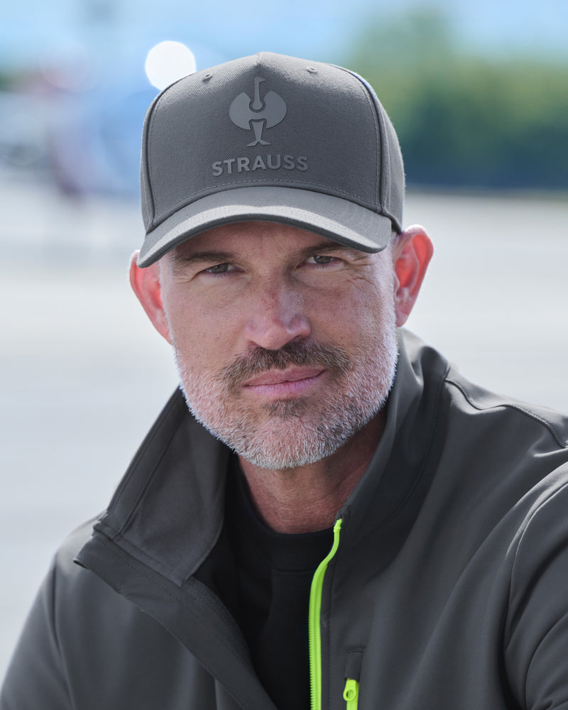 A close-up portrait of a man wearing a dark gray baseball cap with the brand name 'Strauss' and a logo on the front, paired with a dark gray zip-up jacket with a bright neon green zipper.