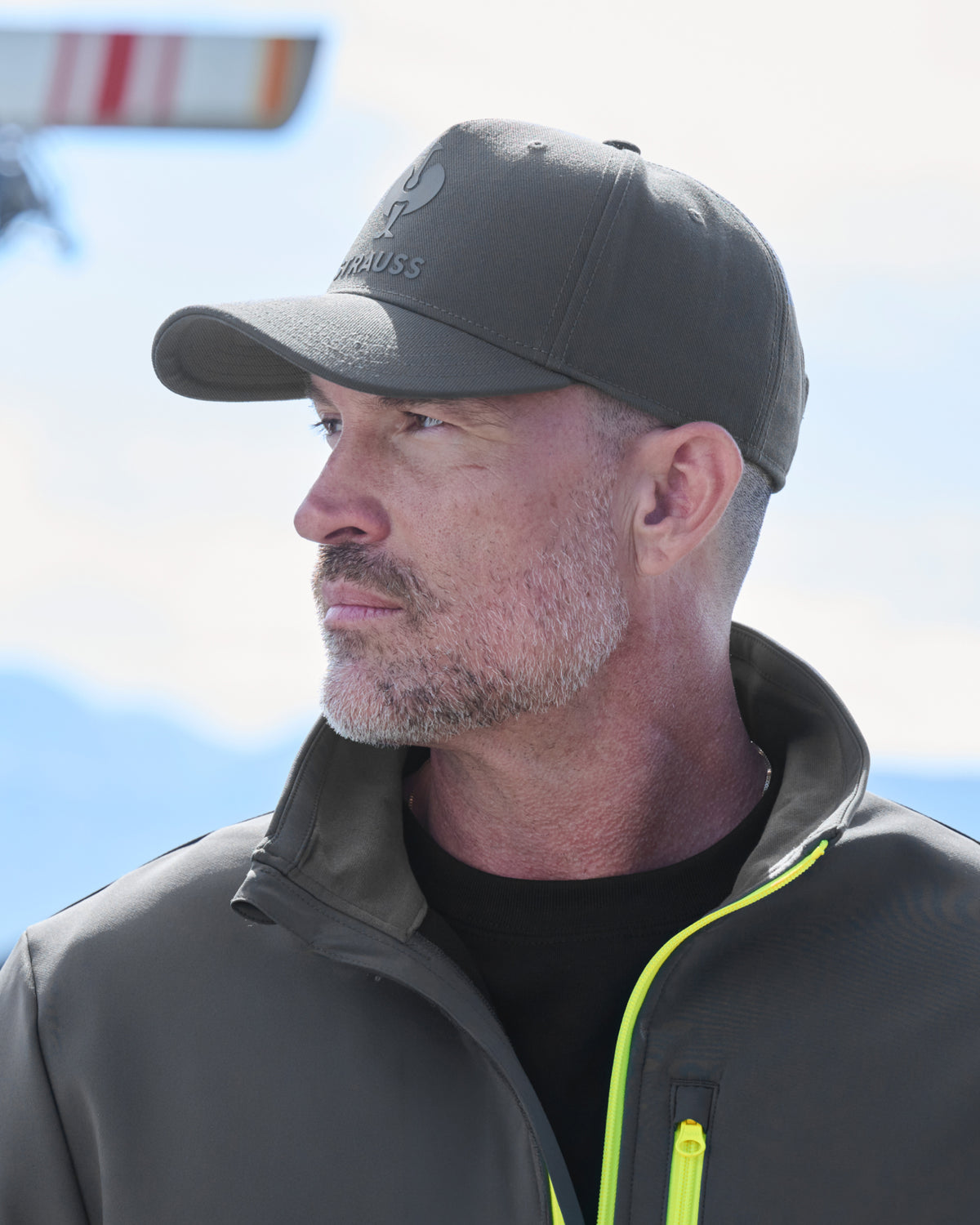 A man in profile wearing a dark gray Ausrüss baseball cap and a dark gray zip-up jacket with a bright neon yellow-green zipper over a black shirt, set against a bright sky background.