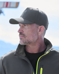 A man in profile wearing a dark gray Ausrüss baseball cap and a dark gray zip-up jacket with a bright neon yellow-green zipper over a black shirt, set against a bright sky background.