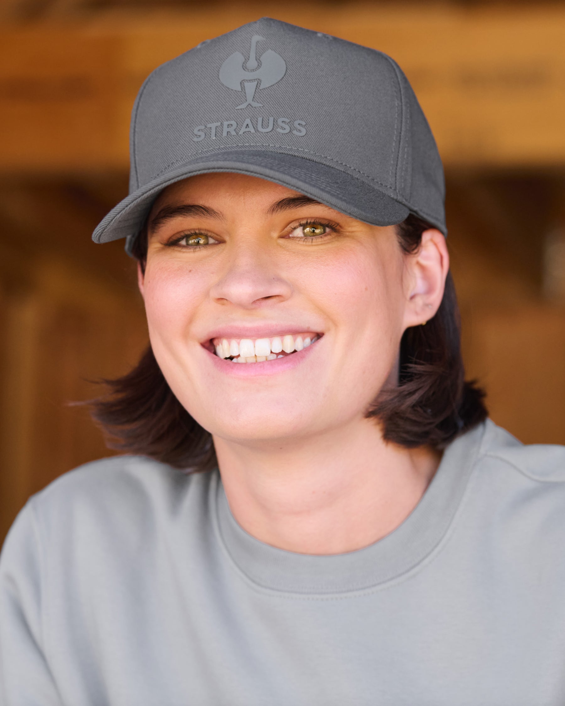 A smiling woman wearing a gray baseball cap with the brand name 'Strauss' and a rooster logo embroidered on the front, paired with a matching gray crewneck sweatshirt against a warm wooden background.