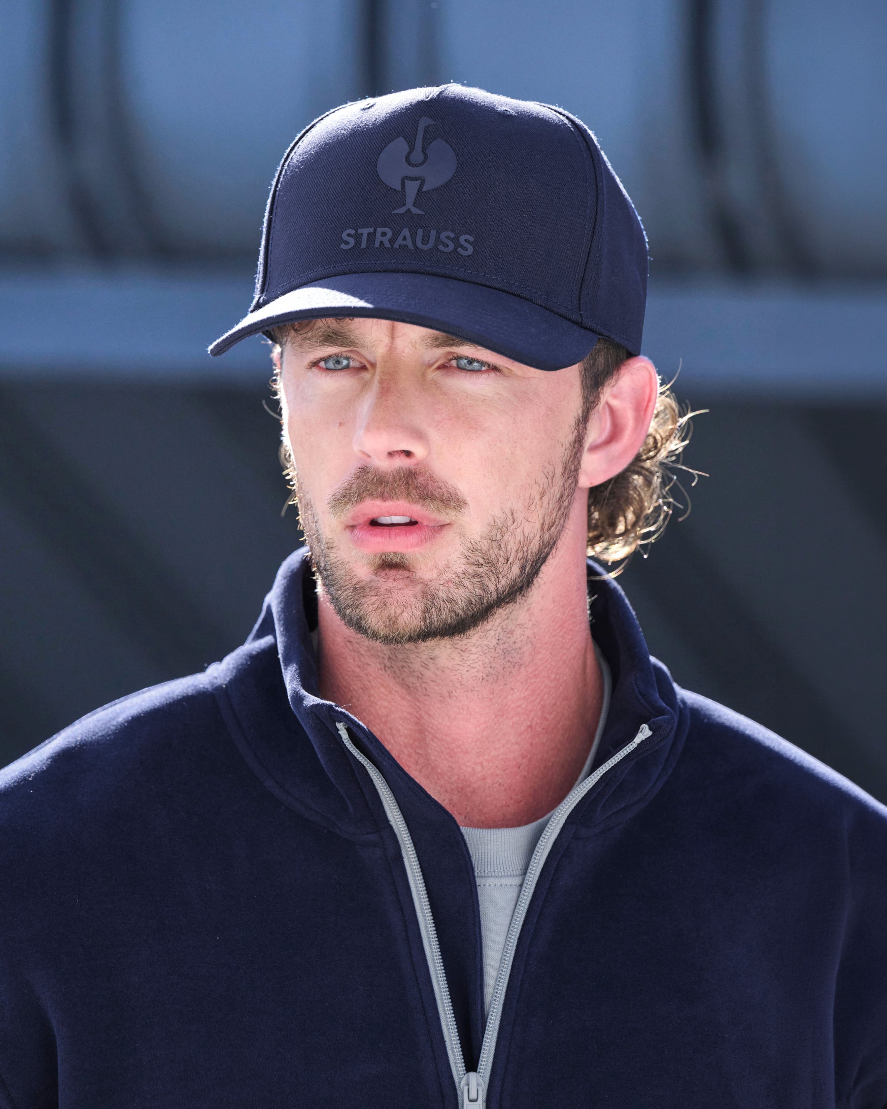 A man wearing a navy blue Strauss baseball cap and a matching navy fleece zip-up jacket over a light gray t-shirt, photographed in bright daylight.