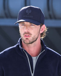 A man wearing a navy blue Strauss baseball cap and a matching navy fleece zip-up jacket over a light gray t-shirt, photographed in bright daylight.