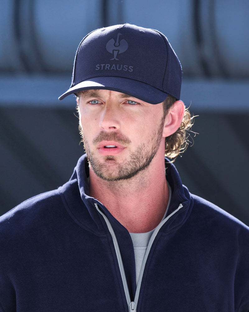 A man wearing a navy blue Strauss baseball cap and a matching navy fleece zip-up jacket over a light gray t-shirt, photographed in bright daylight.