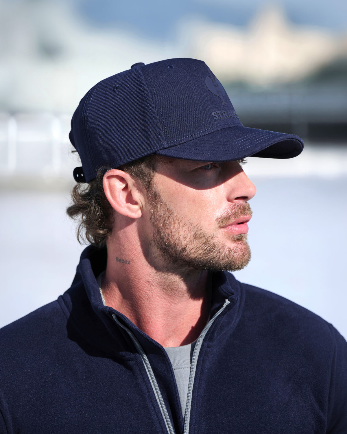 A man in profile wearing a navy blue baseball cap with a subtle embroidered logo and a matching navy fleece quarter-zip pullover, set against a bright, blurred outdoor background.