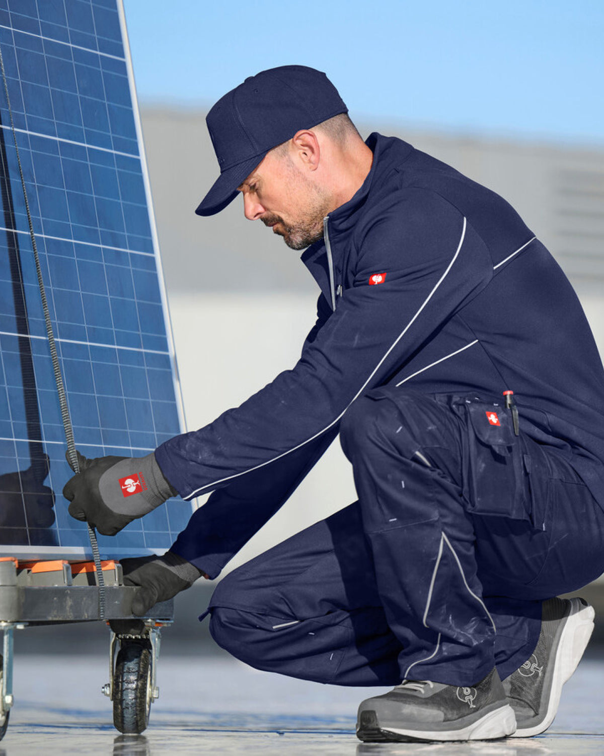 A man in a navy blue work uniform and cap kneels beside a solar panel, handling a cable on a cart in bright outdoor light.