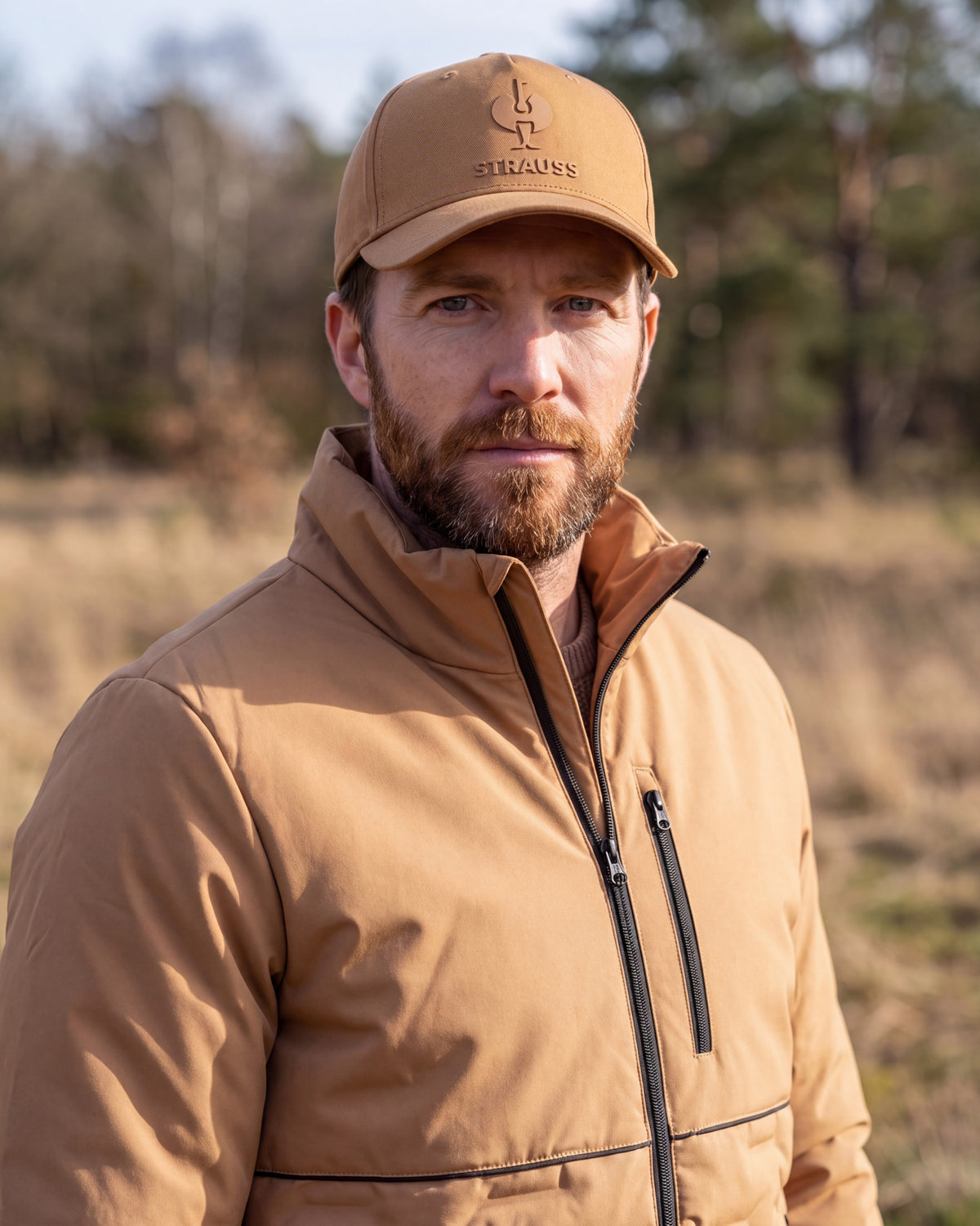 A man wearing a tan Stiegess cap and a matching tan zip-up jacket, photographed outdoors in a natural field setting.