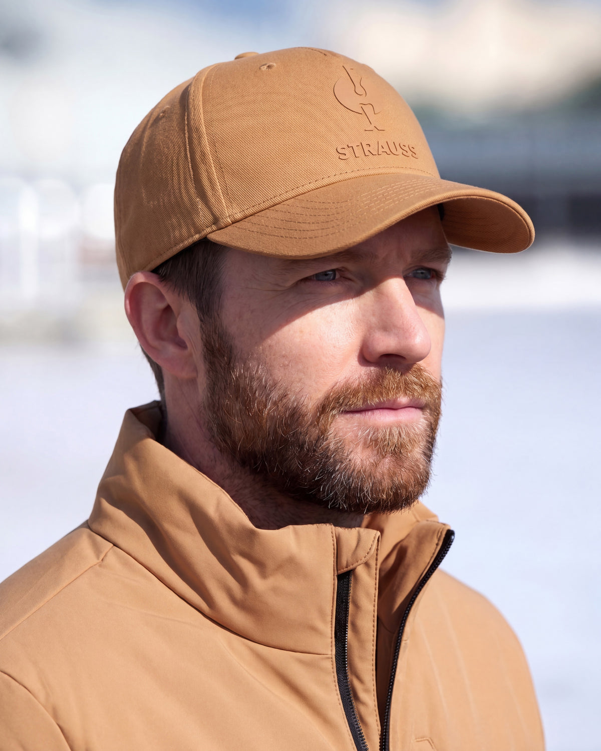 A man wearing a tan STRAUSS baseball cap and a matching tan zip-up jacket, photographed outdoors against a bright, blurred background.