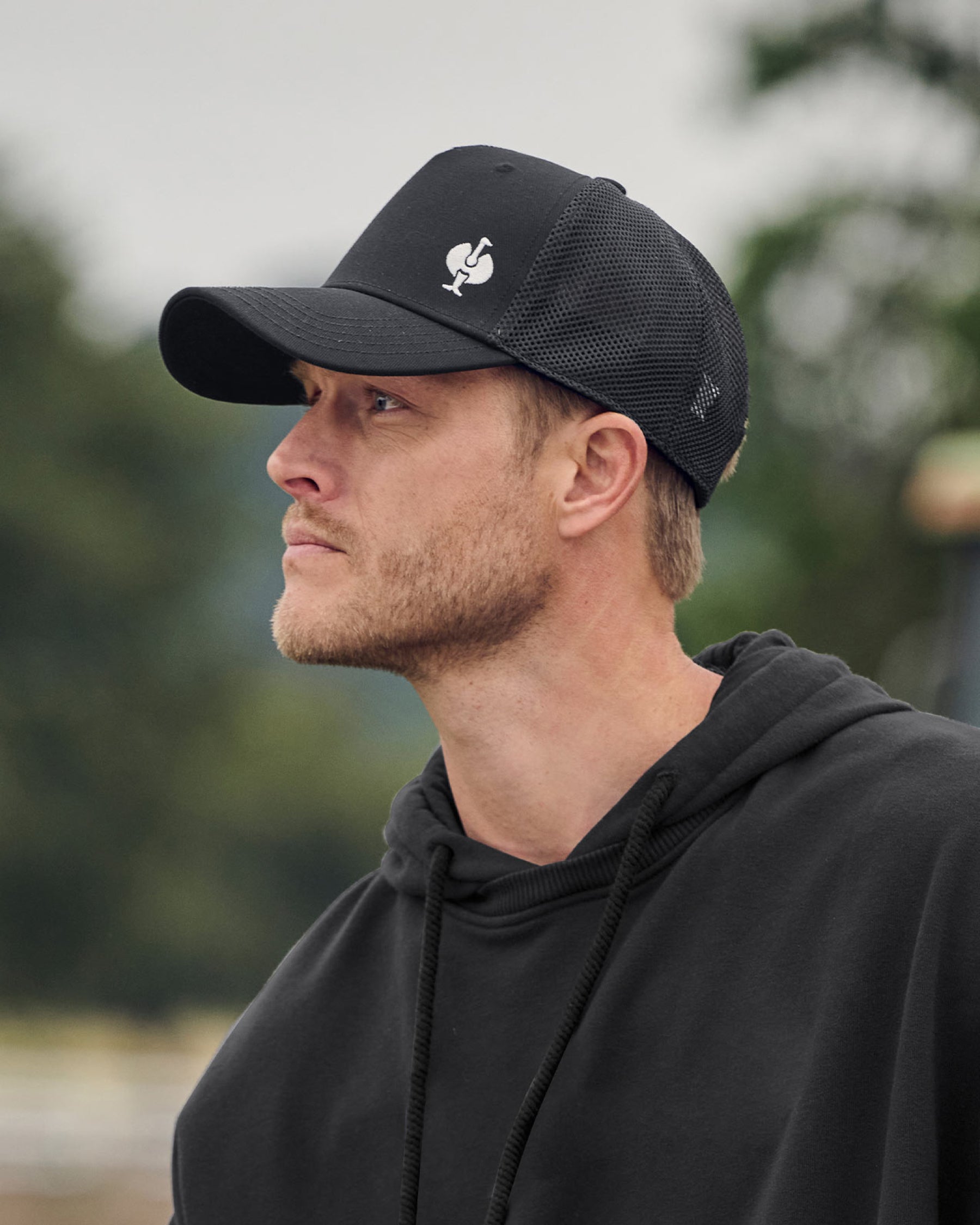 A worker in a black STRAUSS e.s.ventura vintage trucker hat stands near jobsite materials in outdoor workwear.