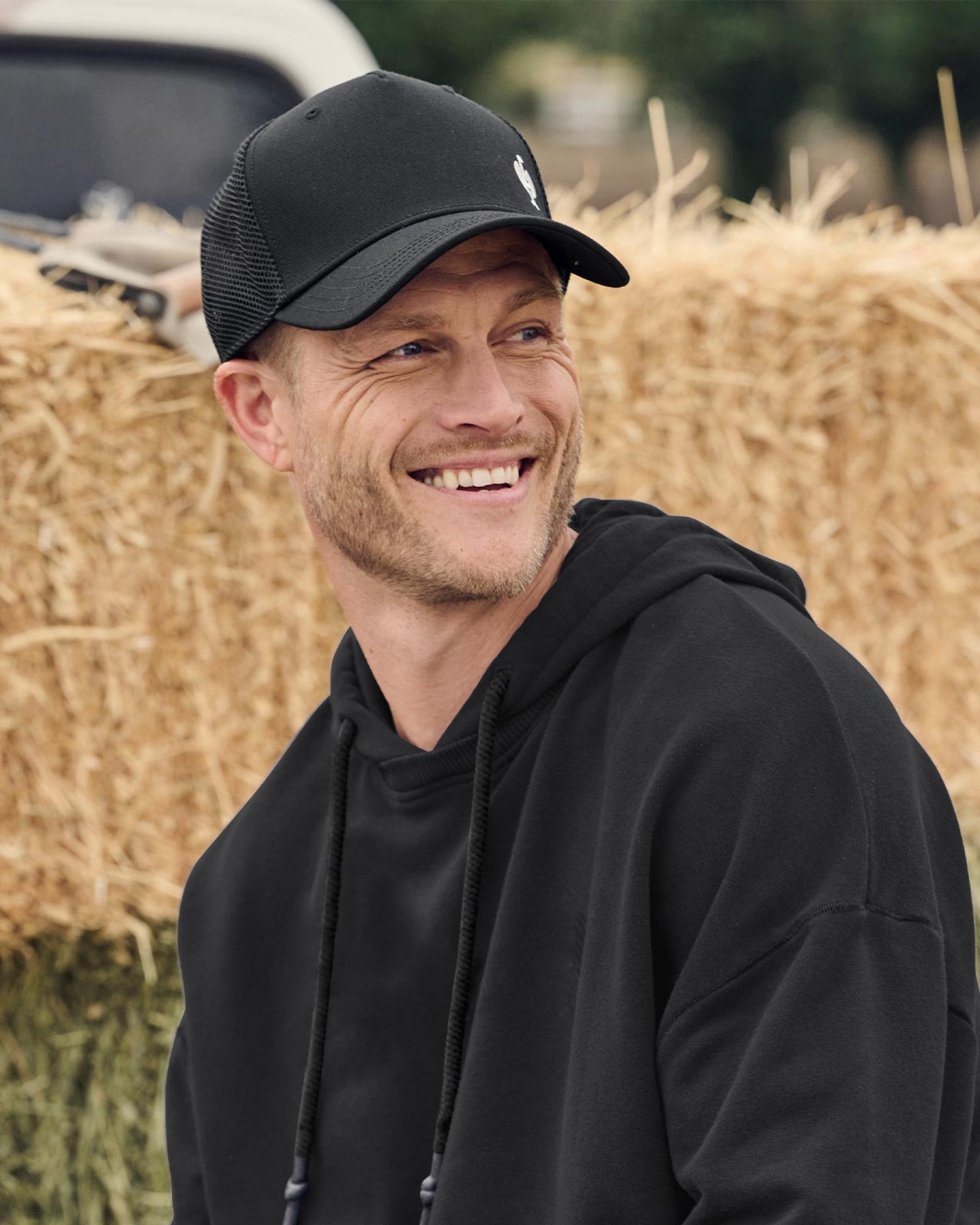 A person wears a black STRAUSS e.s.ventura vintage trucker hat with a jacket and work pants in an outdoor setting.