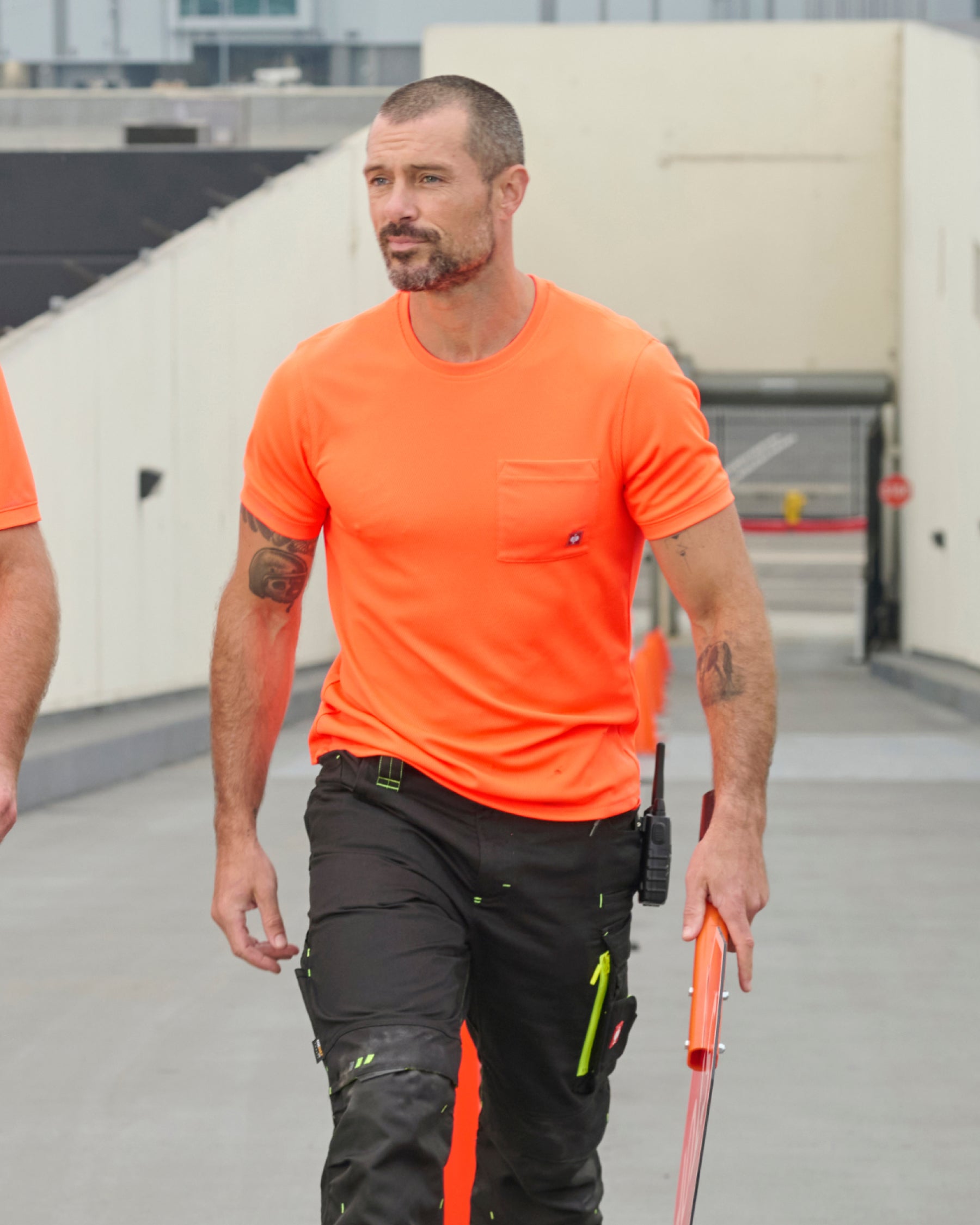 A worker wears the STRAUSS Enhanced Visibility Performance T-Shirt in neon orange with dark work pants near construction equipment