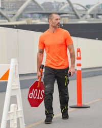 A worker wears the STRAUSS Enhanced Visibility Performance T-Shirt in neon orange with work pants while handling materials at a jobsite