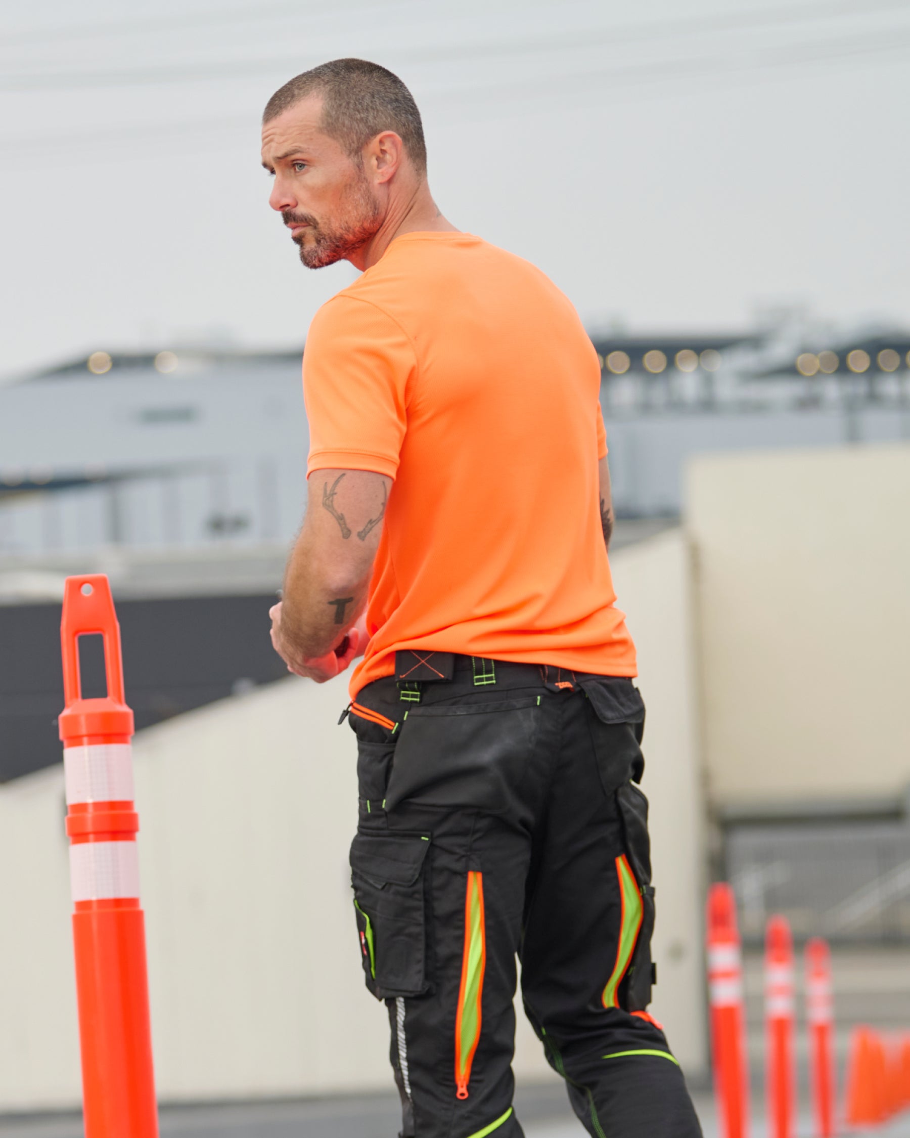 A worker wears the STRAUSS Enhanced Visibility Performance T-Shirt in neon orange with work pants beside a work truck and jobsite gear
