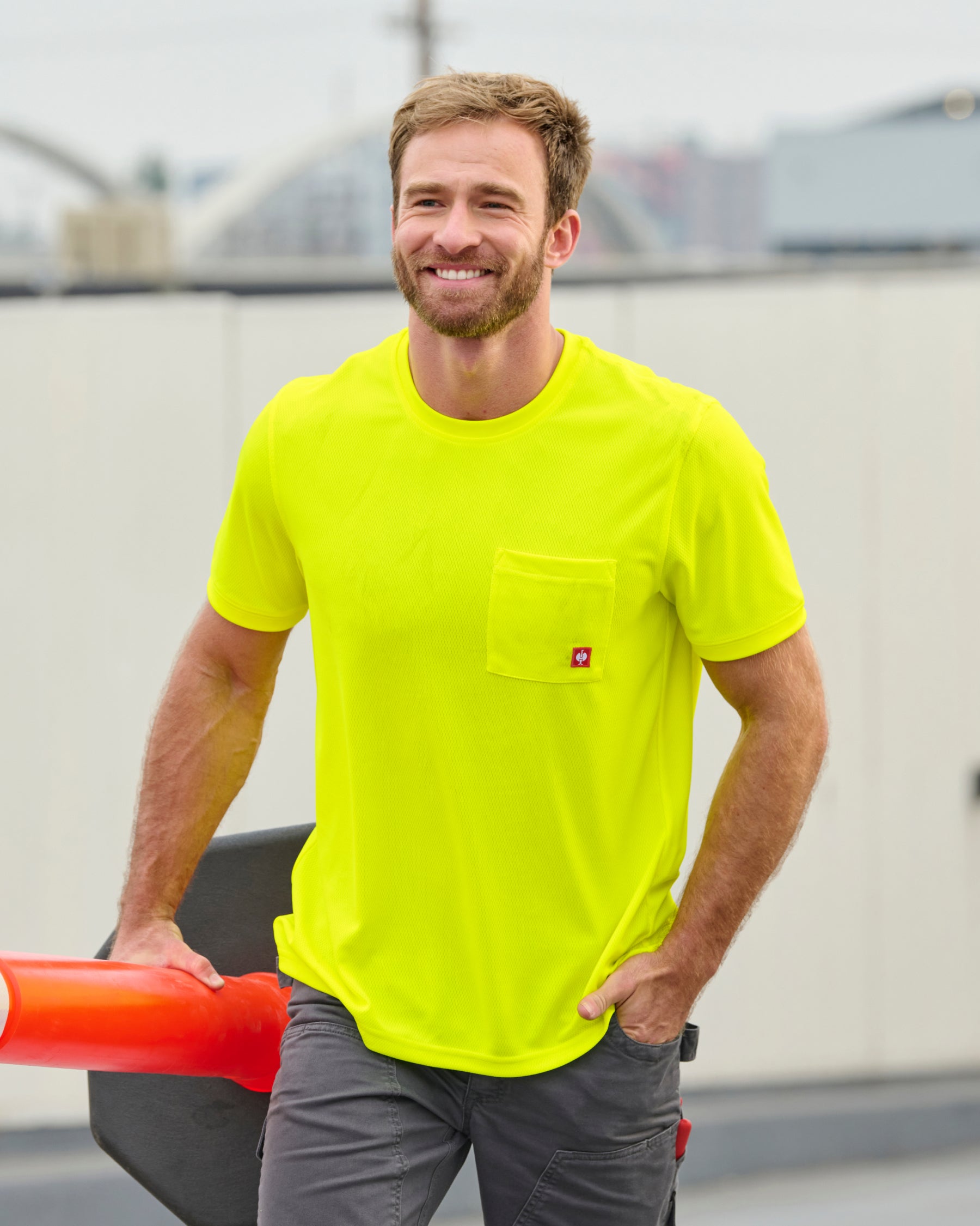 A worker wears the STRAUSS Enhanced Visibility Performance T-Shirt in neon yellow with dark work pants near construction equipment