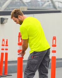 A worker wears the STRAUSS Enhanced Visibility Performance T-Shirt in neon yellow with work pants beside a work truck and jobsite gear