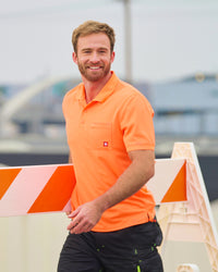 A worker wears the STRAUSS Enhanced Visibility Performance Polo Shirt in neon orange with dark work pants near construction equipment