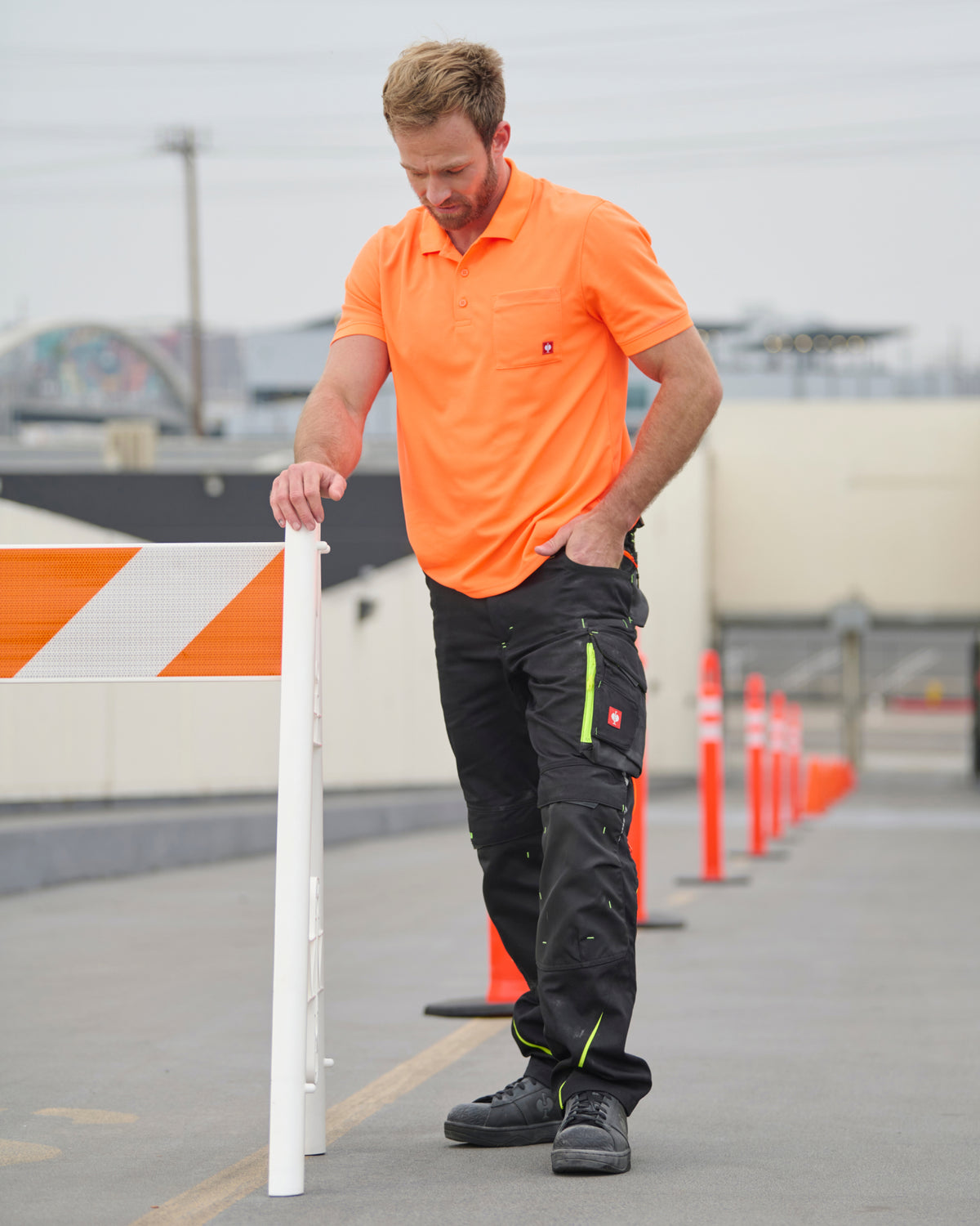 A worker wears the STRAUSS Enhanced Visibility Performance Polo Shirt in neon orange with work pants while handling materials at a jobsite