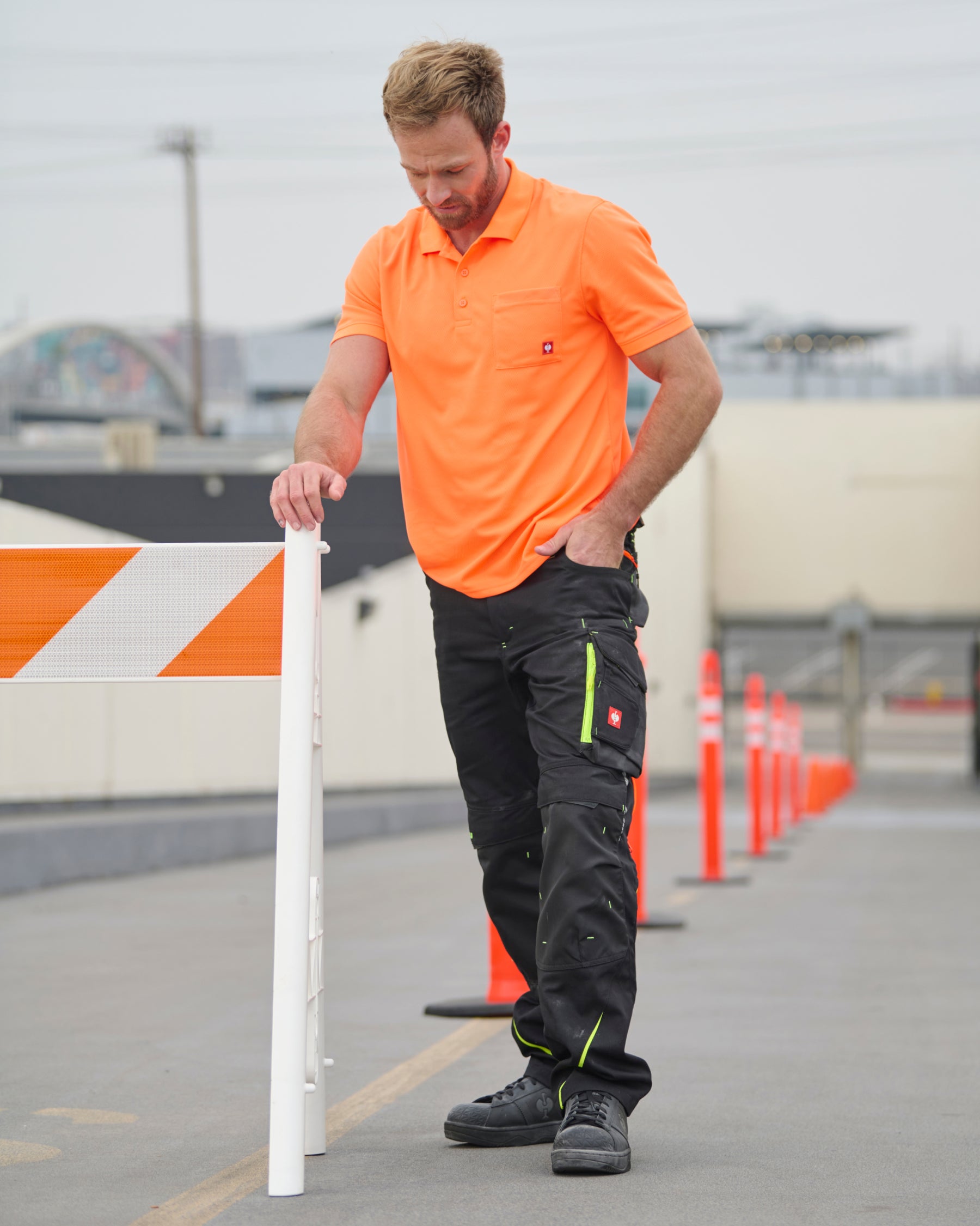 A worker wears the STRAUSS Enhanced Visibility Performance Polo Shirt in neon orange with work pants while handling materials at a jobsite