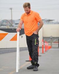 A worker wears the STRAUSS Enhanced Visibility Performance Polo Shirt in neon orange with work pants while handling materials at a jobsite