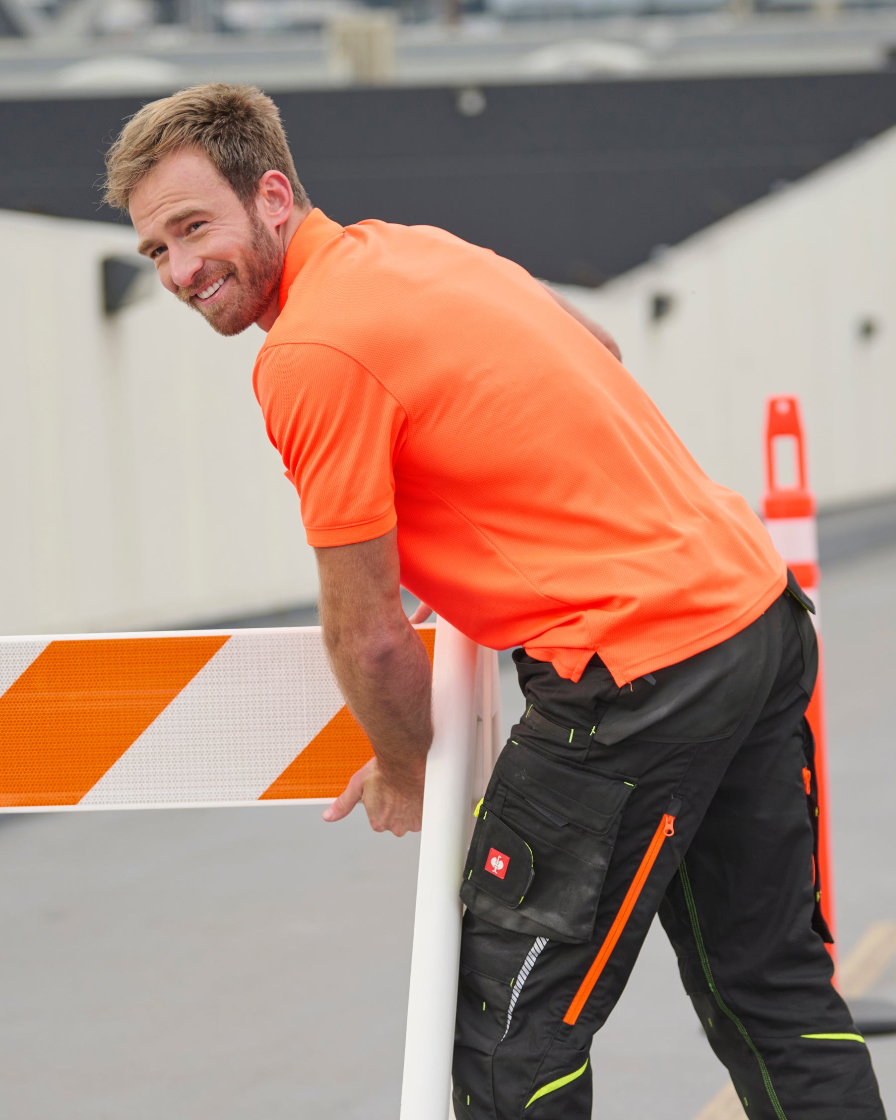 A worker wears the STRAUSS Enhanced Visibility Performance Polo Shirt in neon orange with work pants beside a work truck and jobsite gear