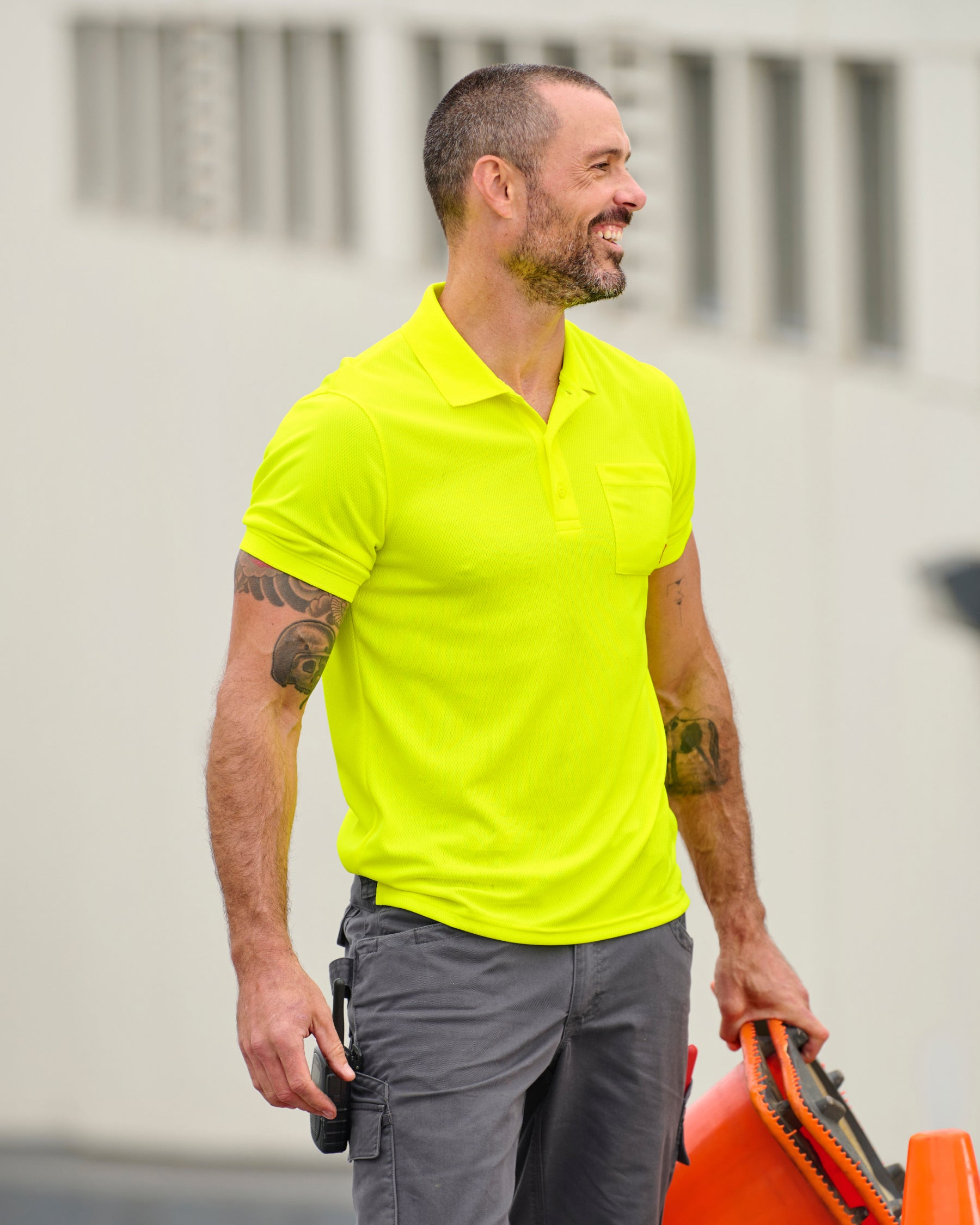 A worker wears the STRAUSS Enhanced Visibility Performance Polo Shirt in neon yellow with work pants beside a work truck and jobsite gear