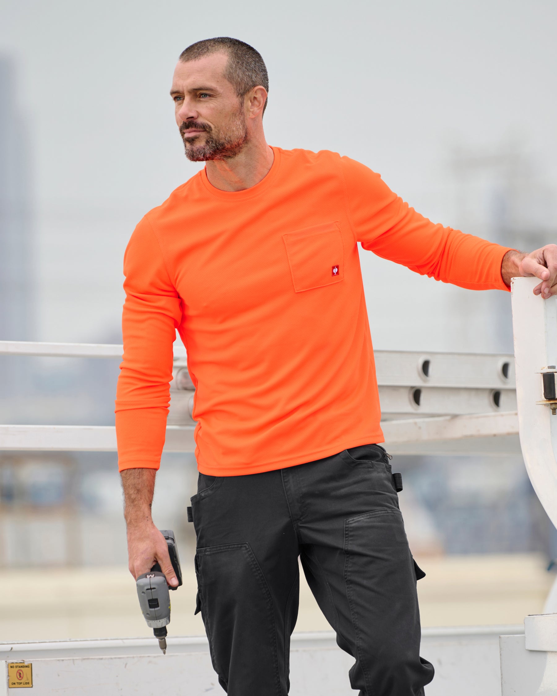 A worker wears the STRAUSS Enhanced Visibility Performance Long Sleeve Shirt in neon orange with dark work pants near construction equipment