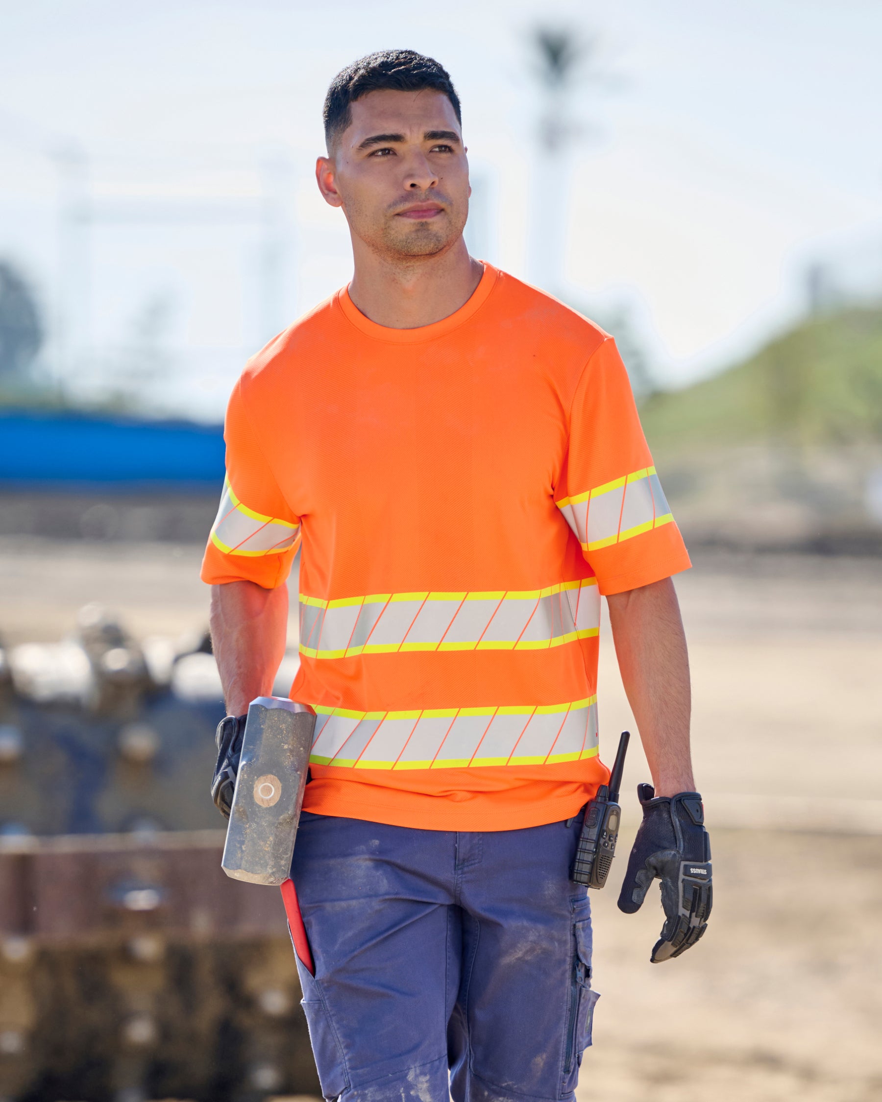 A worker wears the STRAUSS Class 2 High Visibility T-Shirt in neon orange with dark work pants near construction equipment