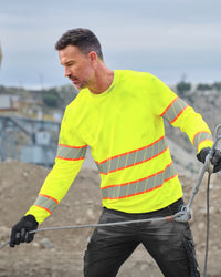 A worker wears the STRAUSS Class 2 High Visibility Long Sleeve Shirt in neon yellow with dark work pants near construction equipment