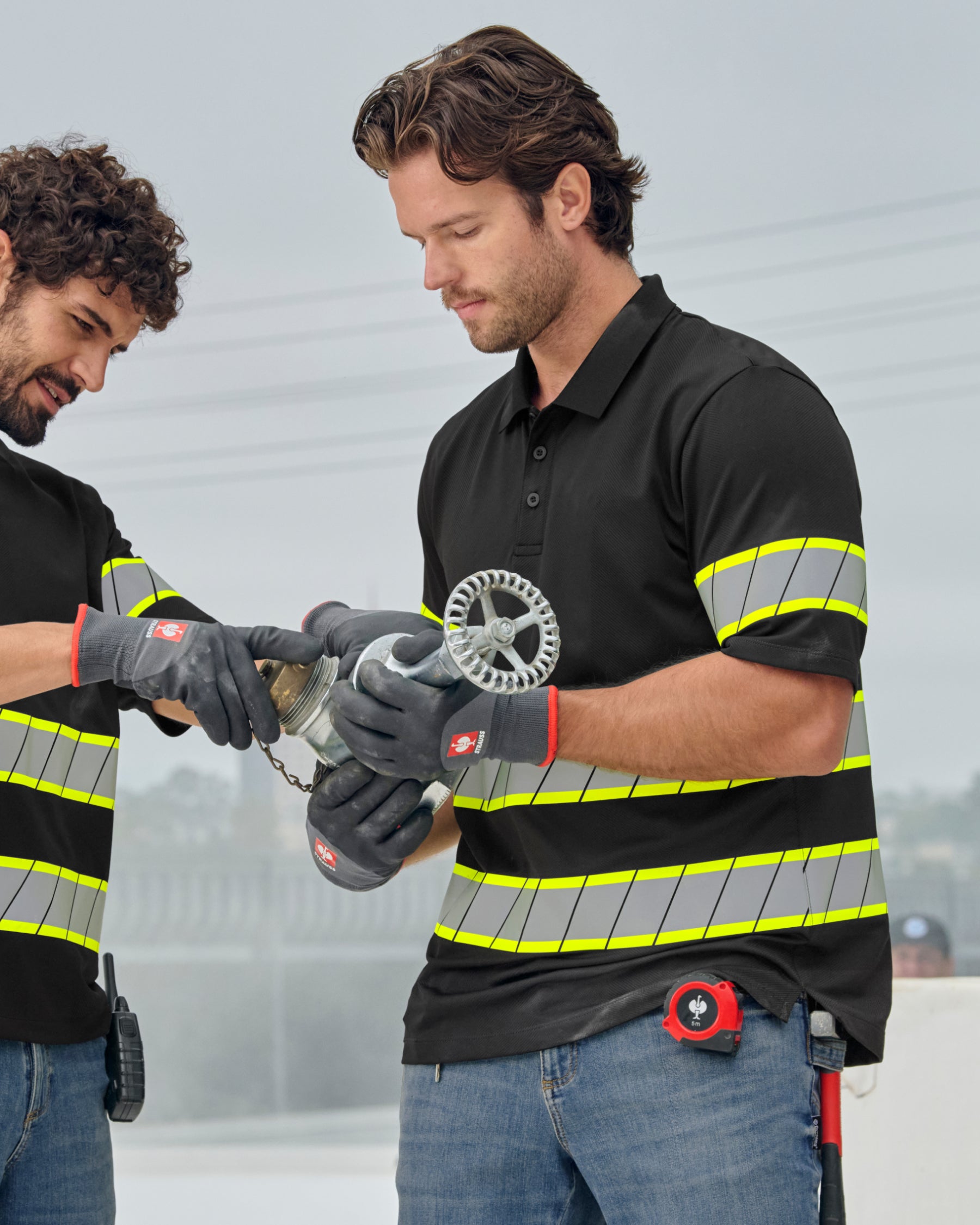 A worker wears the STRAUSS Enhanced Visibility Polo Shirt in black with dark work pants near construction equipment
