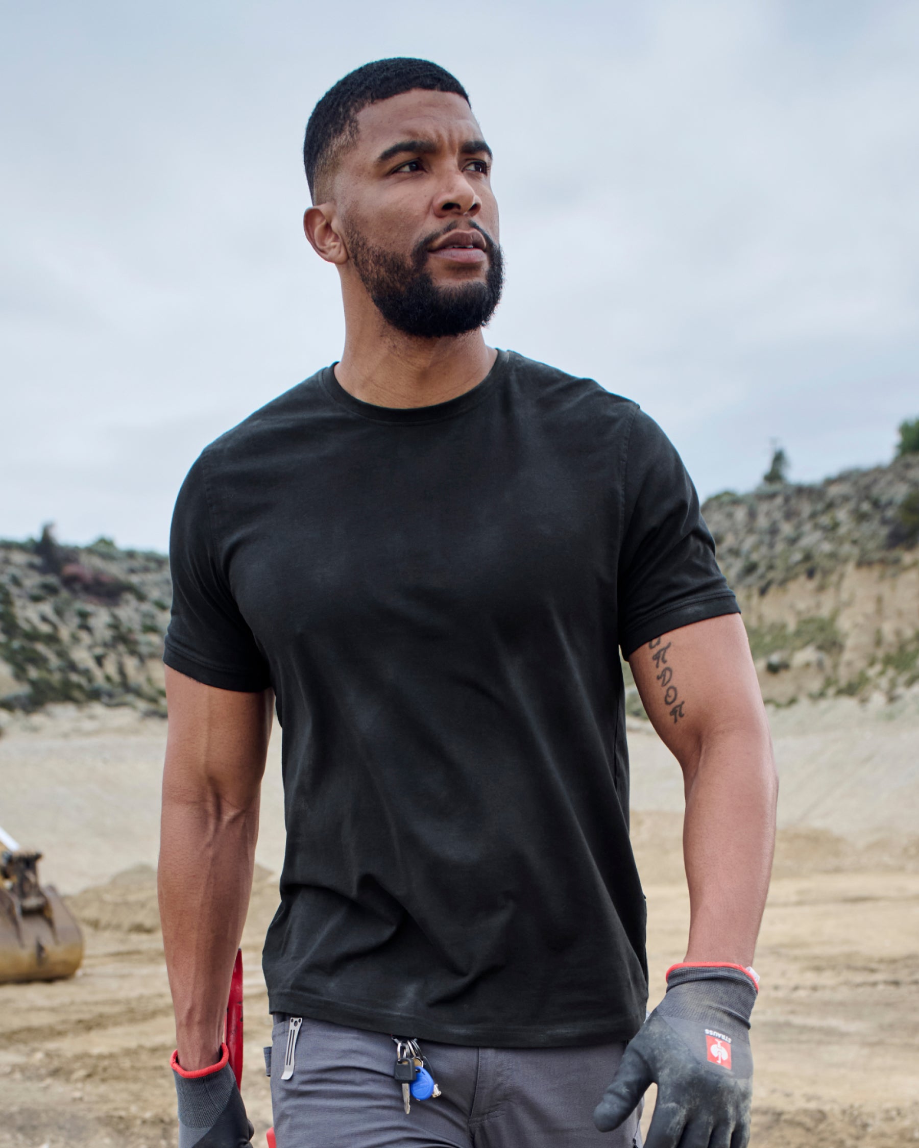 A worker wears the STRAUSS Cotton Stretch T-Shirt in black with tan work pants on a sunny jobsite