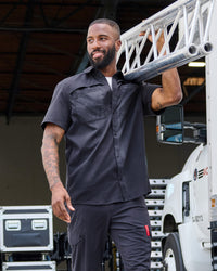 A worker wears the STRAUSS e.s.taktik Stretch Ripstop Short Sleeve Work Shirt in nero blue over a T-shirt with cargo work pants at a jobsite