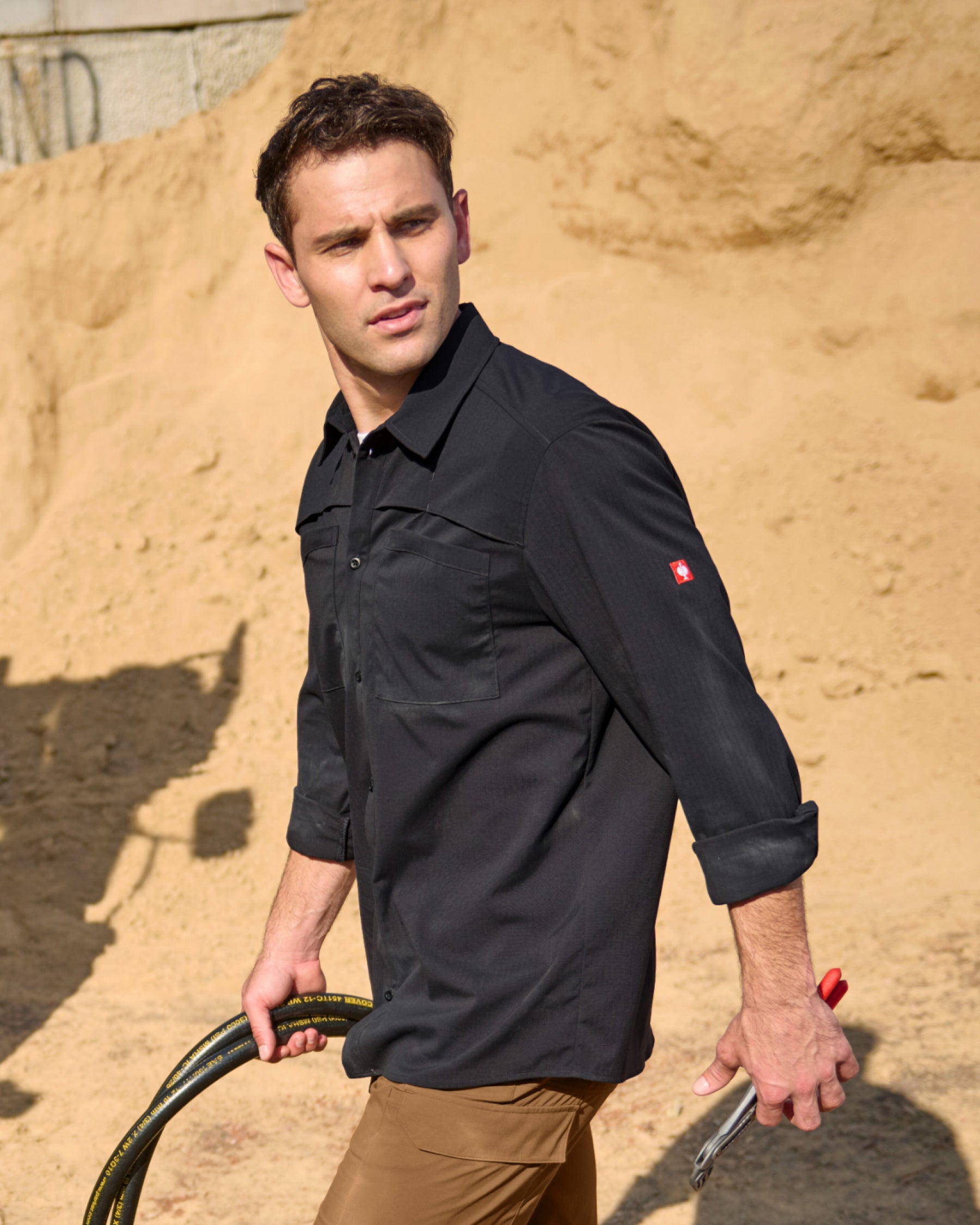 A worker wears the STRAUSS e.s.taktik Stretch Ripstop Long Sleeve Work Shirt in black over a T-shirt with cargo work pants at a jobsite