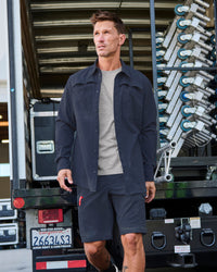 A worker wears the STRAUSS e.s.taktik Stretch Ripstop Long Sleeve Work Shirt in nero blue over a T-shirt with cargo work pants at a jobsite
