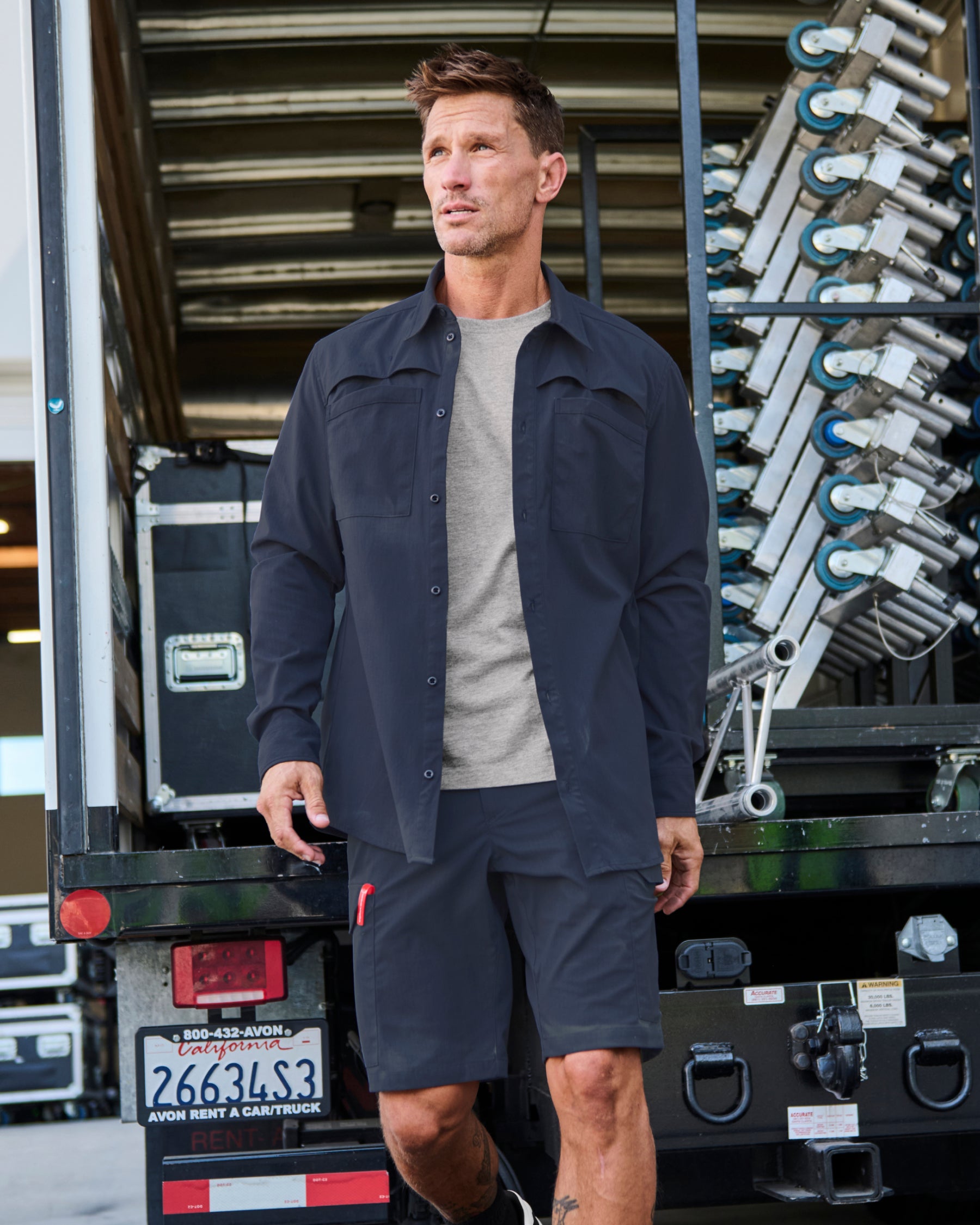 A worker wears the STRAUSS e.s.taktik Stretch Ripstop Long Sleeve Work Shirt in nero blue over a T-shirt with cargo work pants at a jobsite