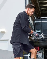 A worker wears the STRAUSS e.s.taktik Stretch Ripstop Long Sleeve Work Shirt in nero blue while moving materials beside metal shelving