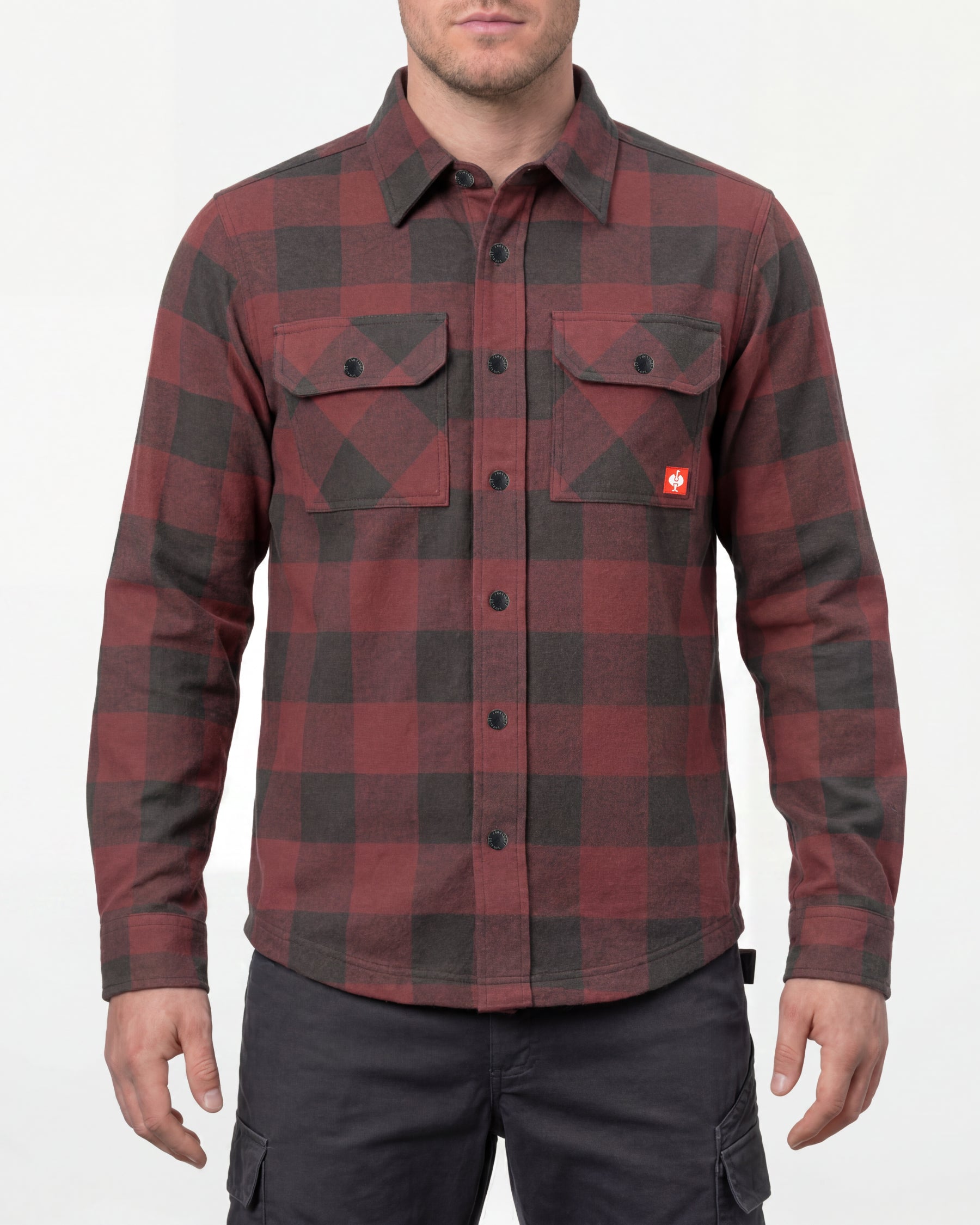 A model wears the STRAUSS e.s.iconic Flannel Work Shirt in oxid red and carbon gray over a T-shirt with work pants and work boots in a studio product scene