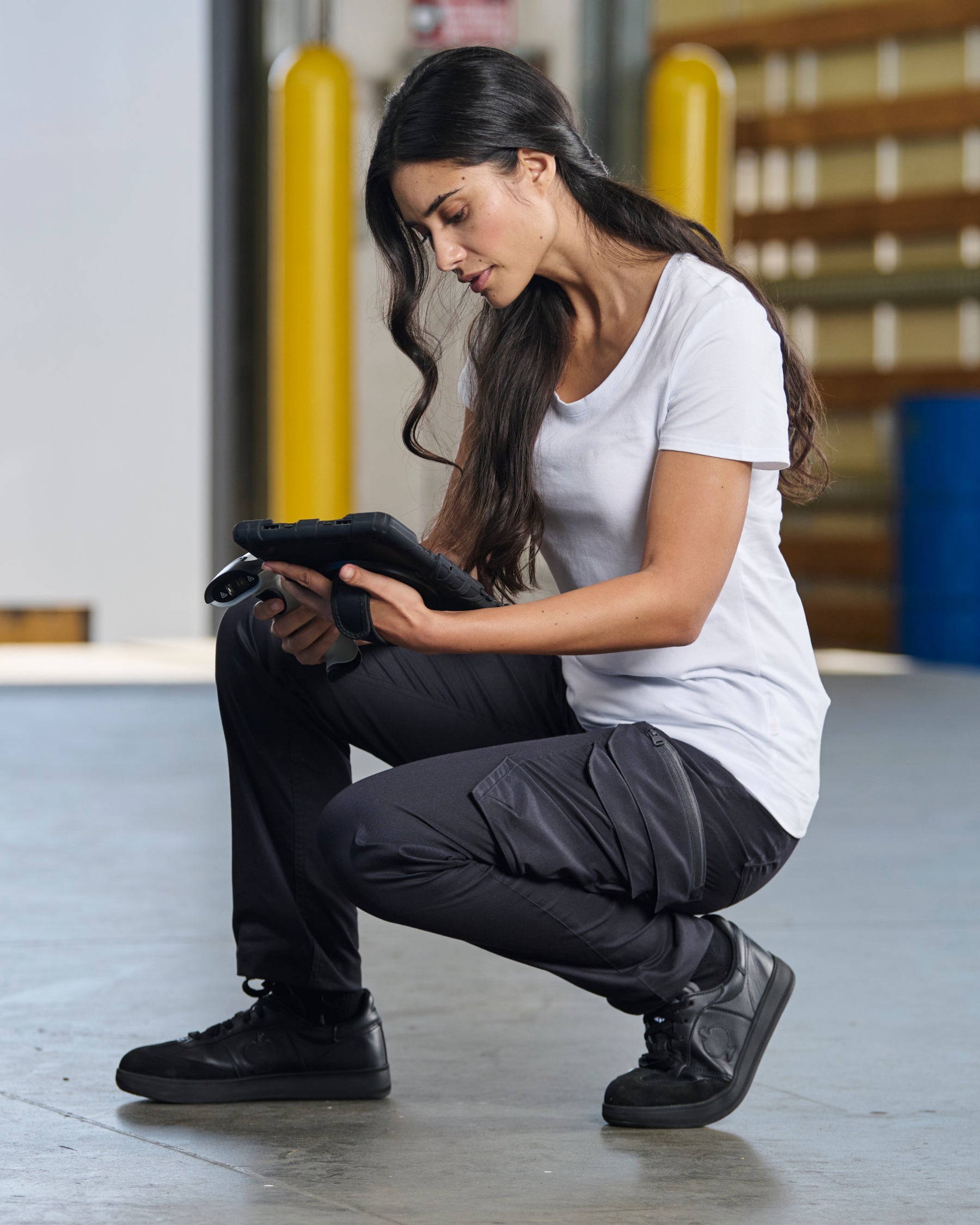 A woman wears the Women's STRAUSS Cotton Stretch T-Shirt in white with work pants beside jobsite equipment