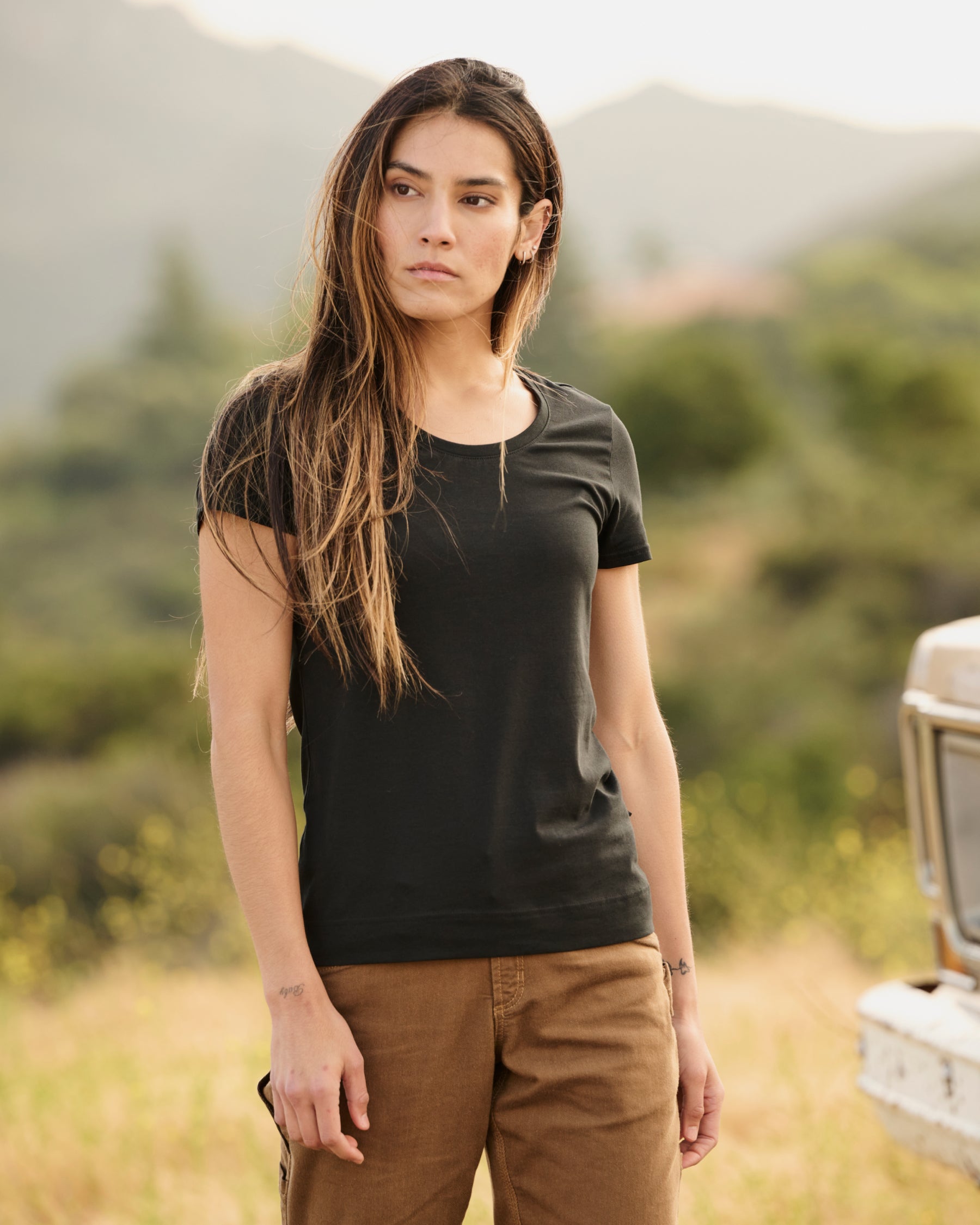 A woman wears the Women's STRAUSS Cotton Stretch T-Shirt in black with work pants on a sunny outdoor workday
