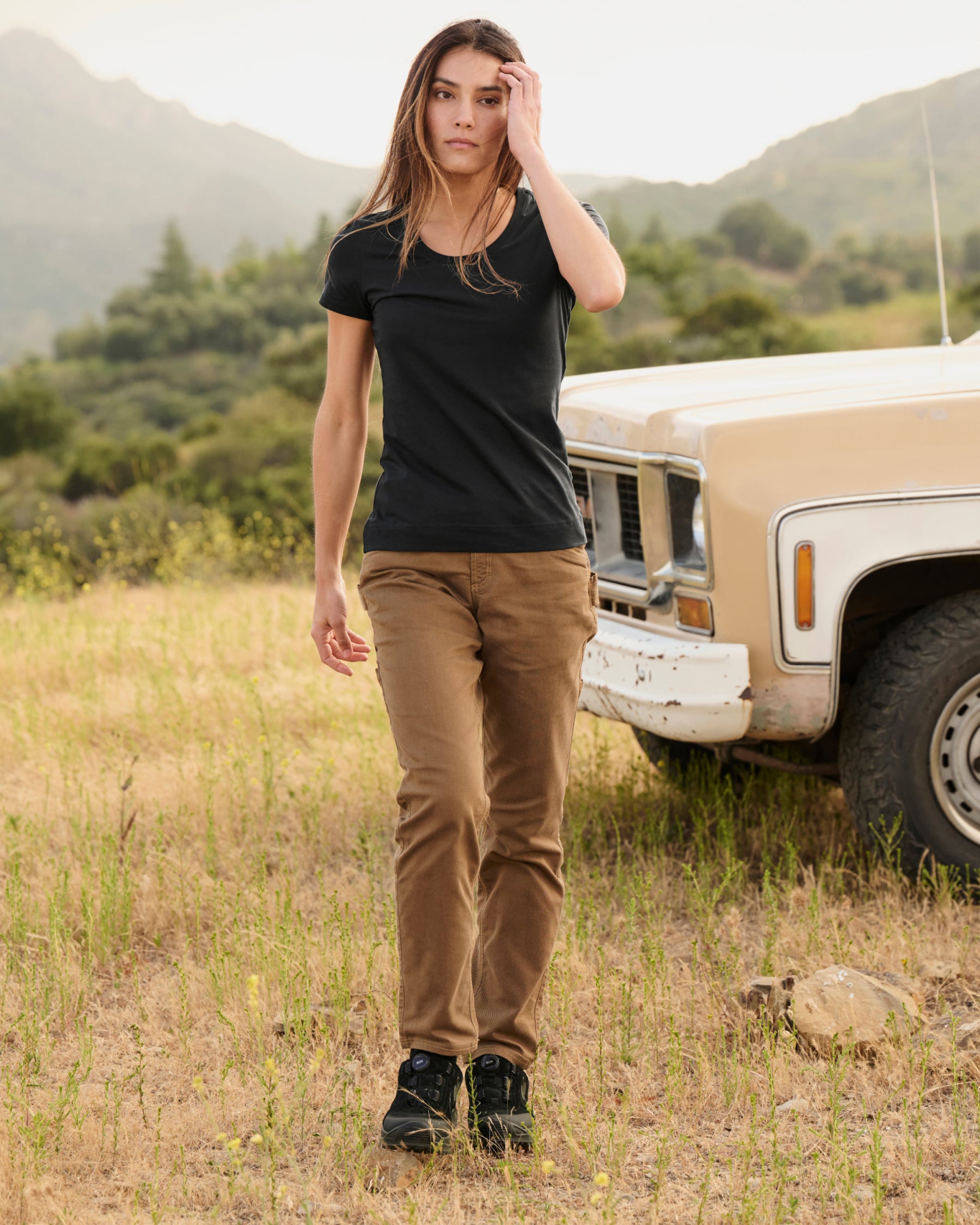 A woman wears the Women's STRAUSS Cotton Stretch T-Shirt in black with work pants beside a field