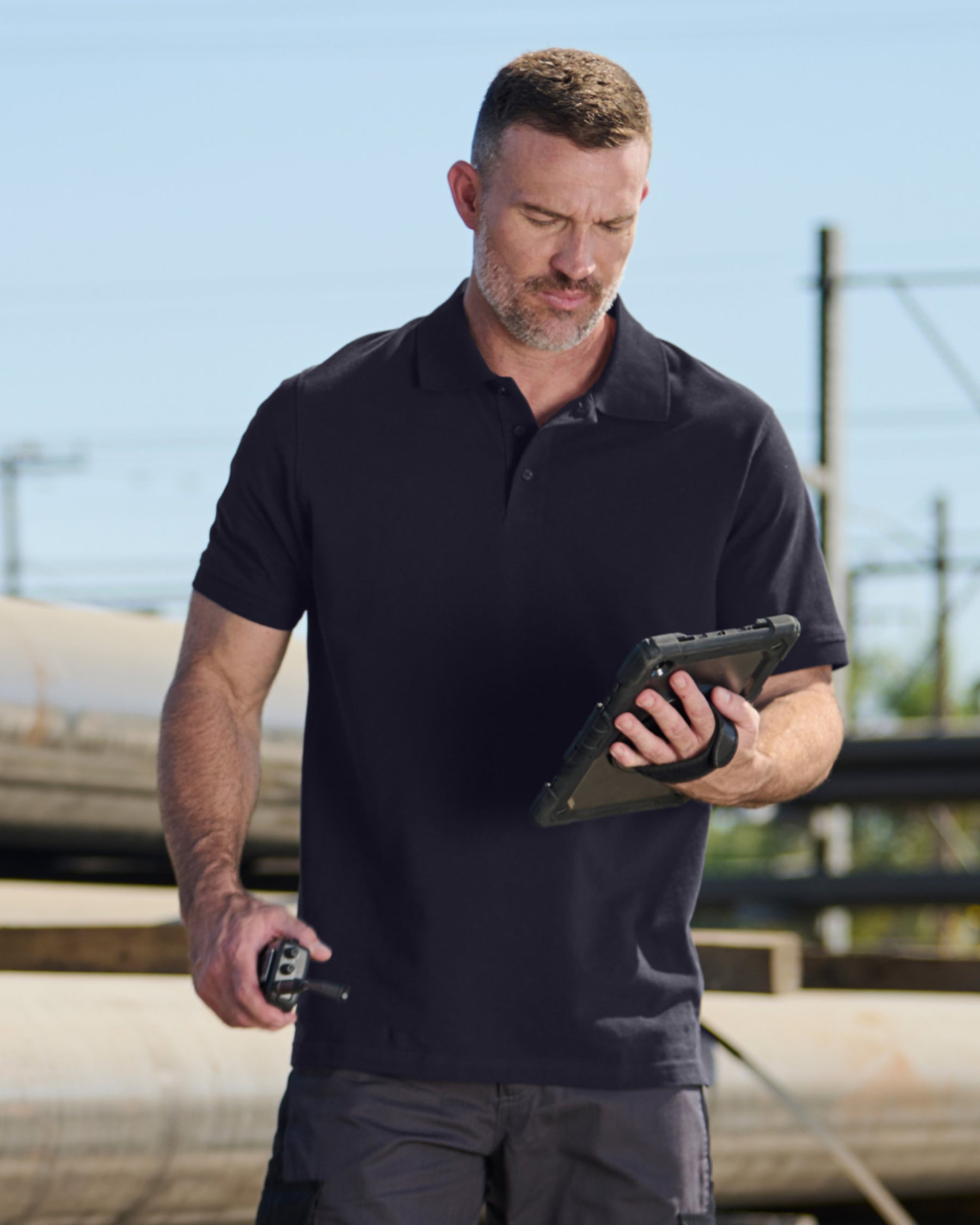 A worker wears the STRAUSS Cotton Polo Shirt in nero blue with tan work pants beside jobsite equipment