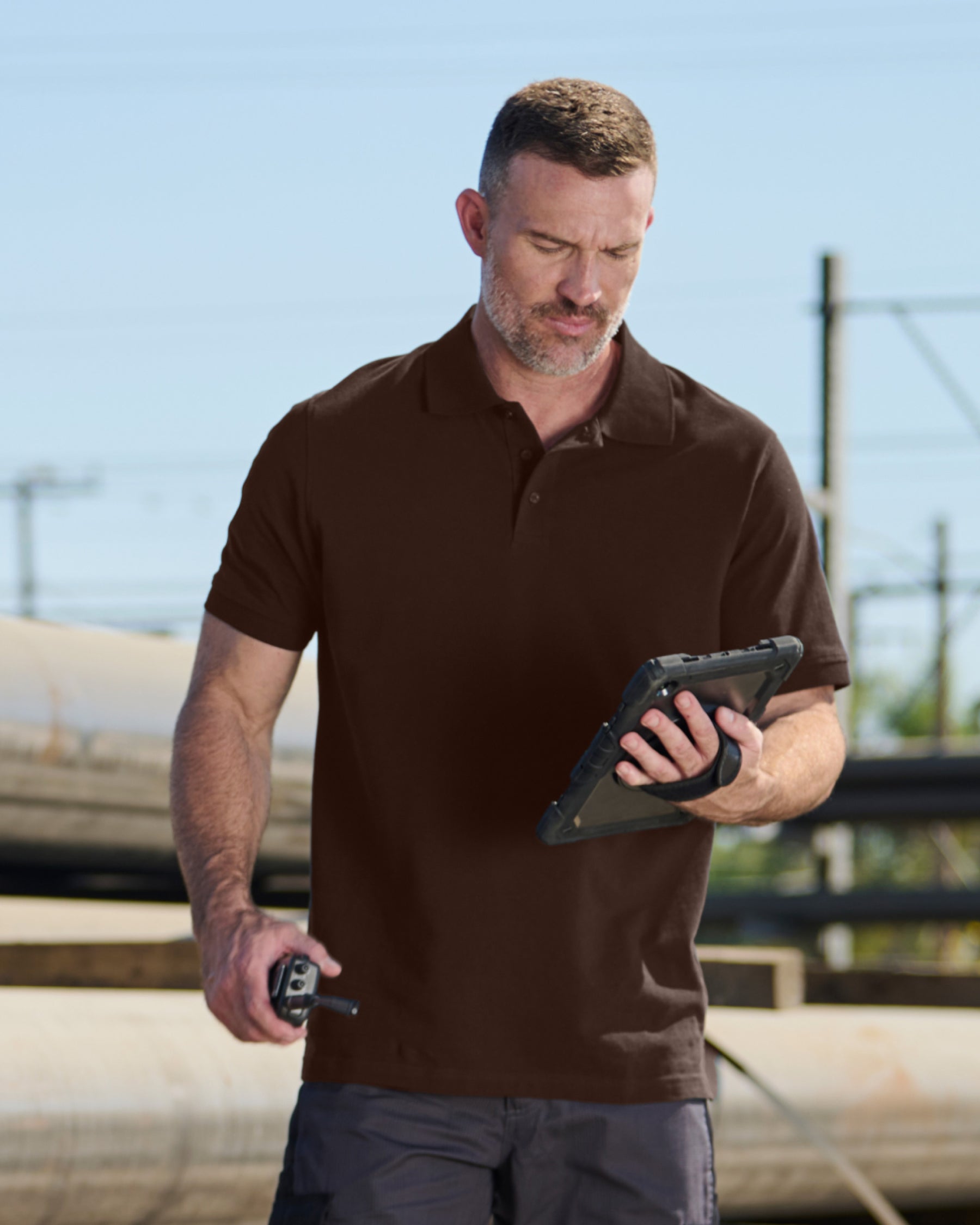 A worker wears the STRAUSS Cotton Polo Shirt in hazelnut with tan work pants beside jobsite equipment