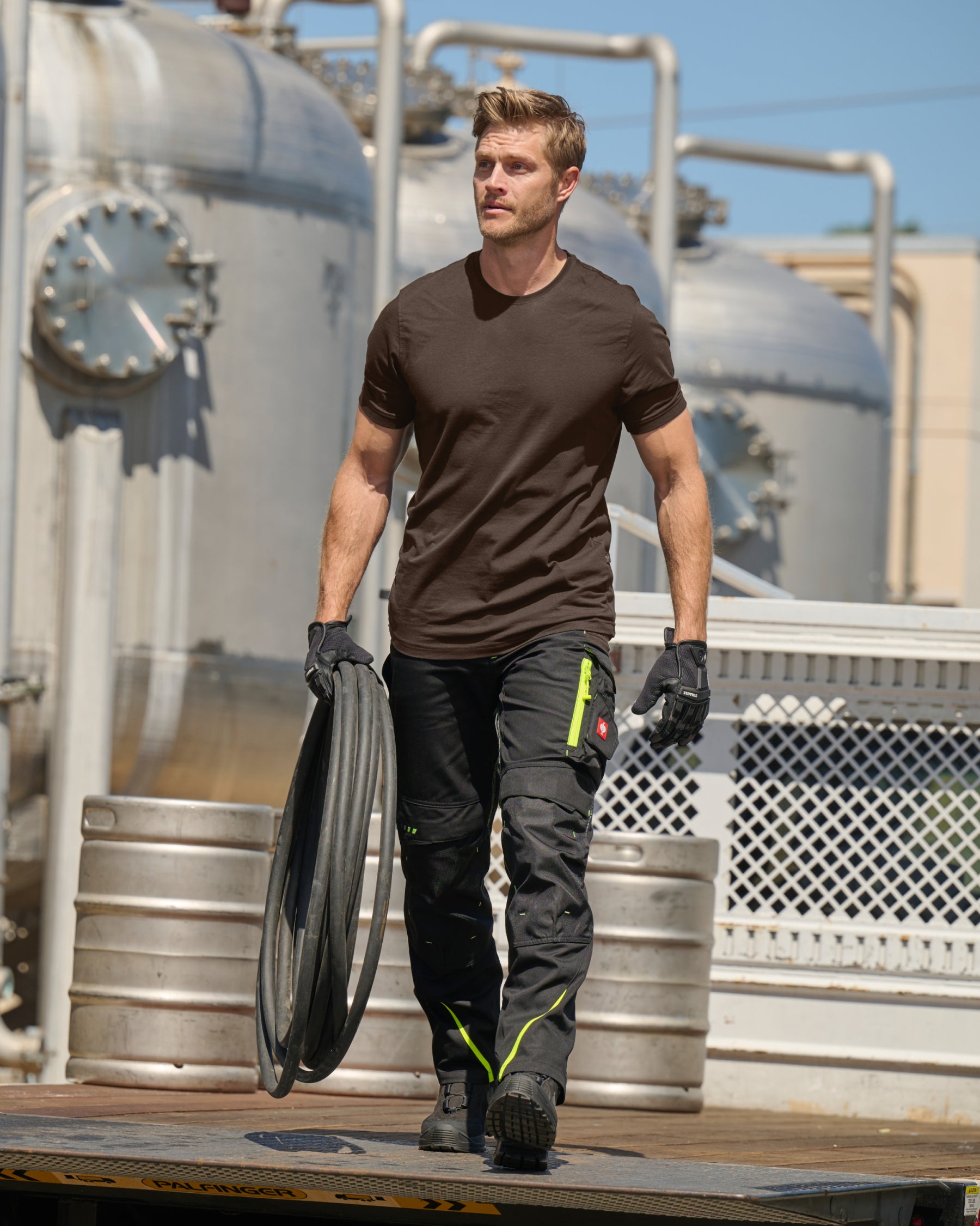 A worker wears the STRAUSS Cotton T-Shirt in hazelnut with tan work pants beside jobsite equipment