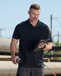 A worker wears the STRAUSS Cotton Polo Shirt in black with tan work pants beside jobsite equipment