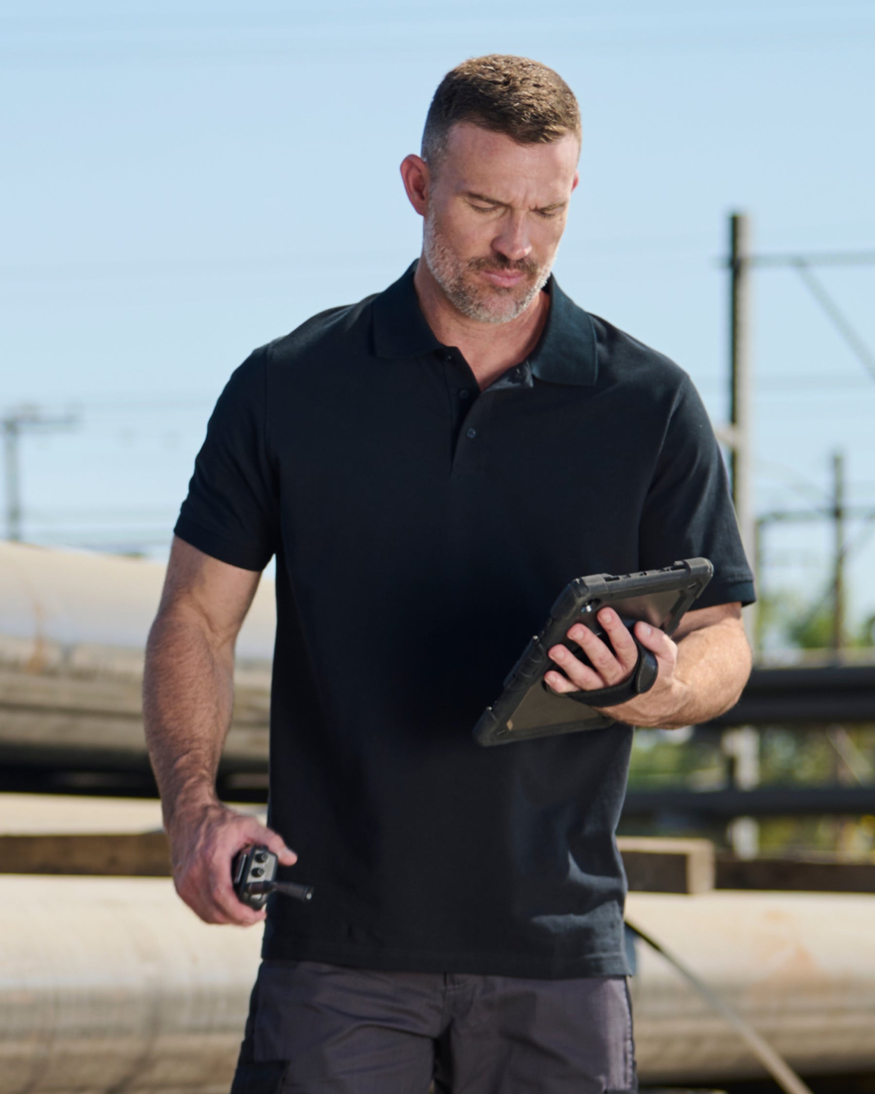 A worker wears the STRAUSS Cotton Polo Shirt in black with tan work pants beside jobsite equipment