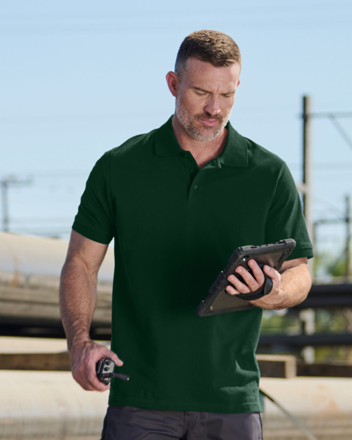 A worker wears the STRAUSS Cotton Polo Shirt in green with tan work pants on a sunny jobsite