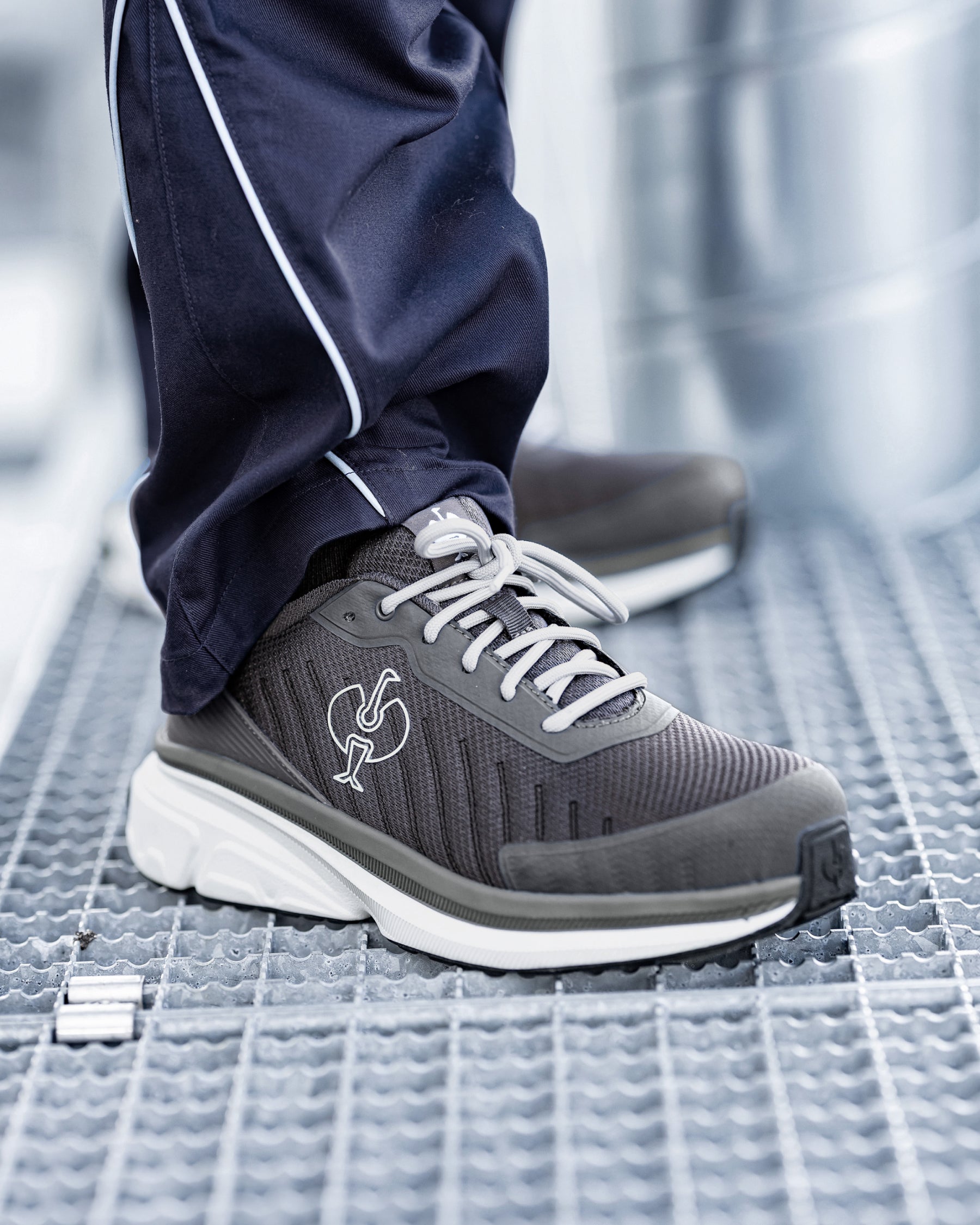 A close-up of a person's foot wearing a dark gray and white athletic sneaker with navy blue pants featuring a white stripe, standing on a textured metal grate.