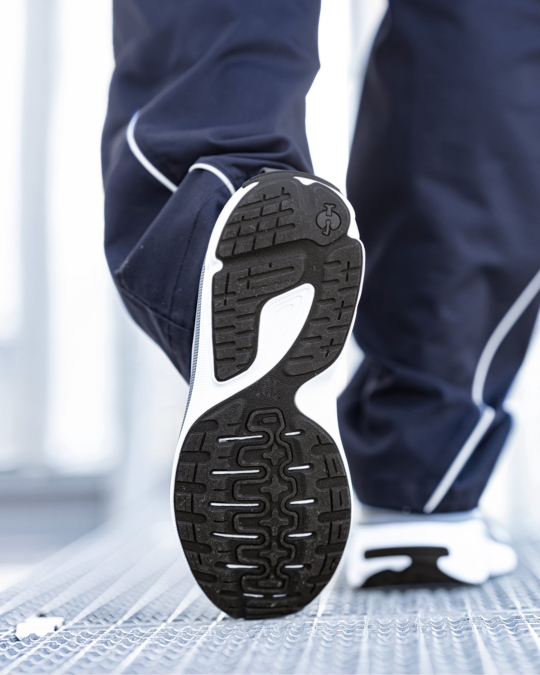 Close-up of a person's leg wearing navy blue athletic pants with white piping and a black-and-white running shoe, showing the textured outsole of the shoe.