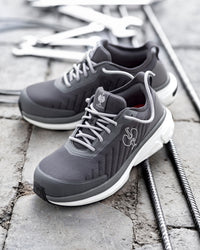 A pair of dark gray and white athletic sneakers with white laces, displayed on a gray stone surface with metal rebar and wrenches in the background.