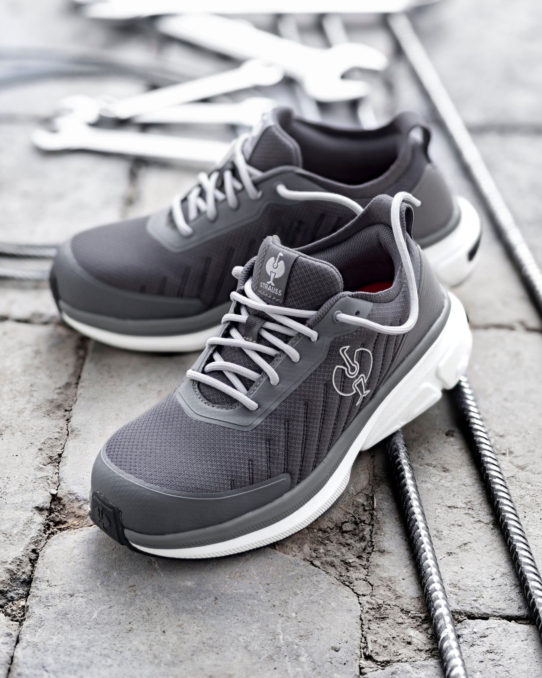 A pair of dark gray and white athletic sneakers with white laces, displayed on a gray stone surface with metal rebar and wrenches in the background.