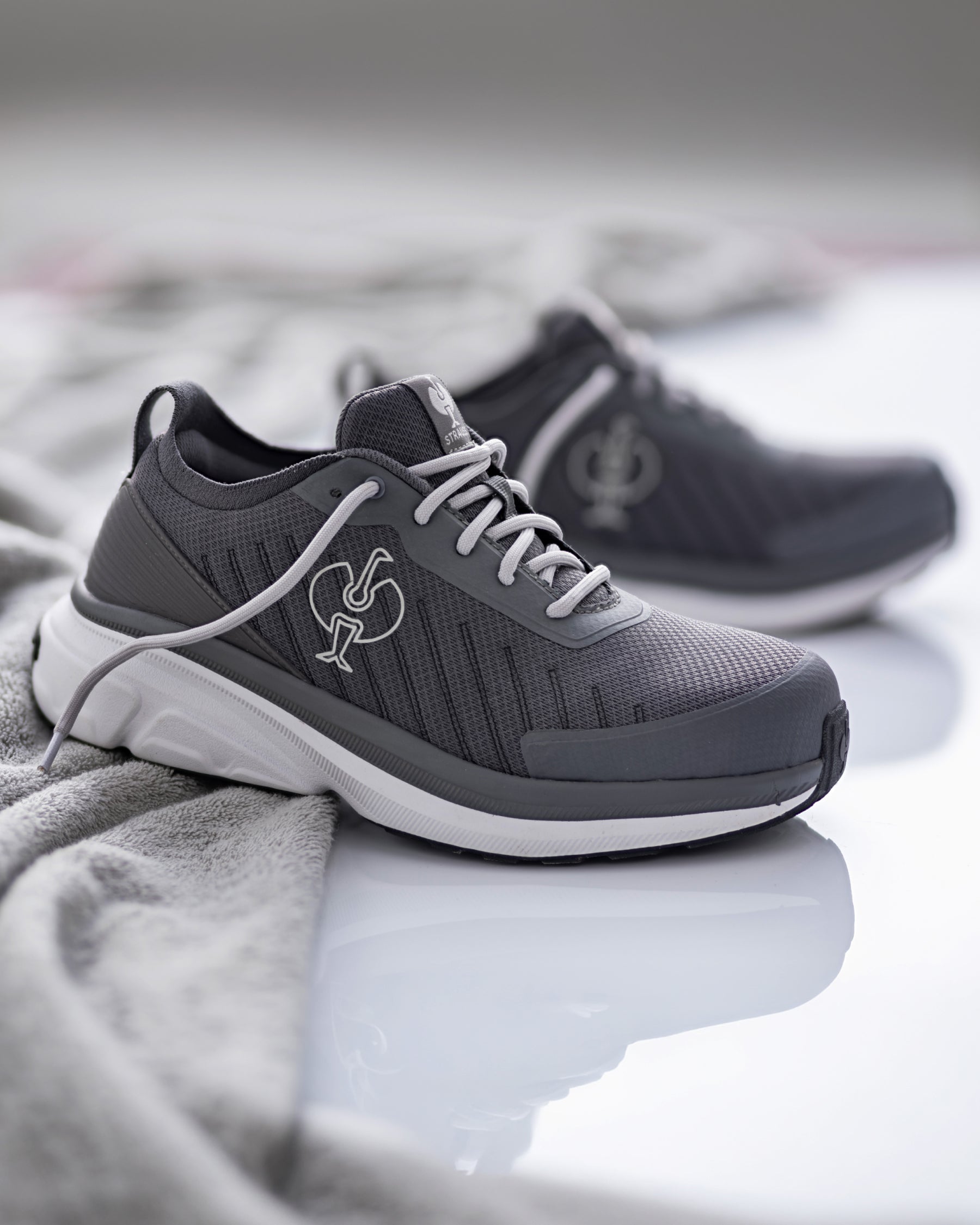 A pair of dark gray athletic sneakers with white laces and a white midsole, displayed on a reflective white surface with a soft gray textured fabric in the foreground.