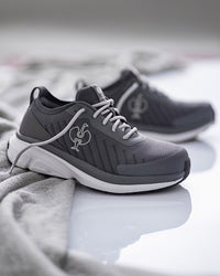 A pair of dark gray athletic sneakers with white laces and a white midsole, displayed on a reflective white surface with a soft gray textured fabric in the foreground.